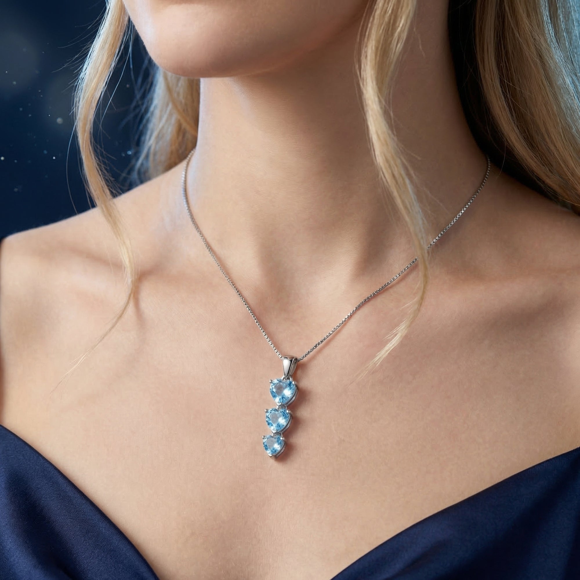 Heart cut blue topaz trio pendant in silver worn by a model in a dark blue dress.