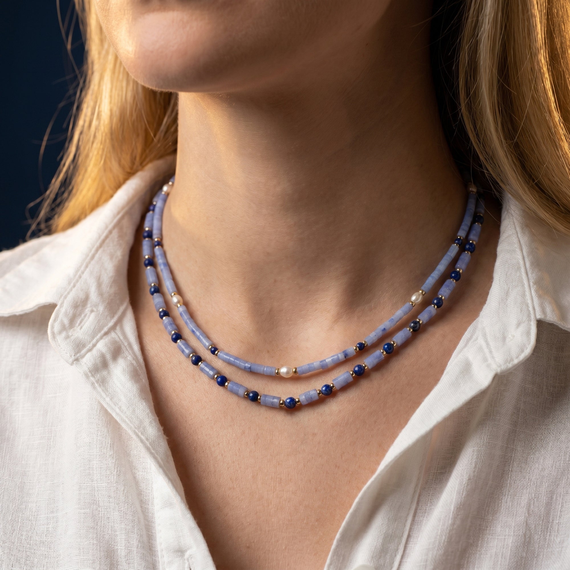 Gold plated layered necklace with cylindrical light blue beads, lapis, and pearls worn by a model in a white blouse.