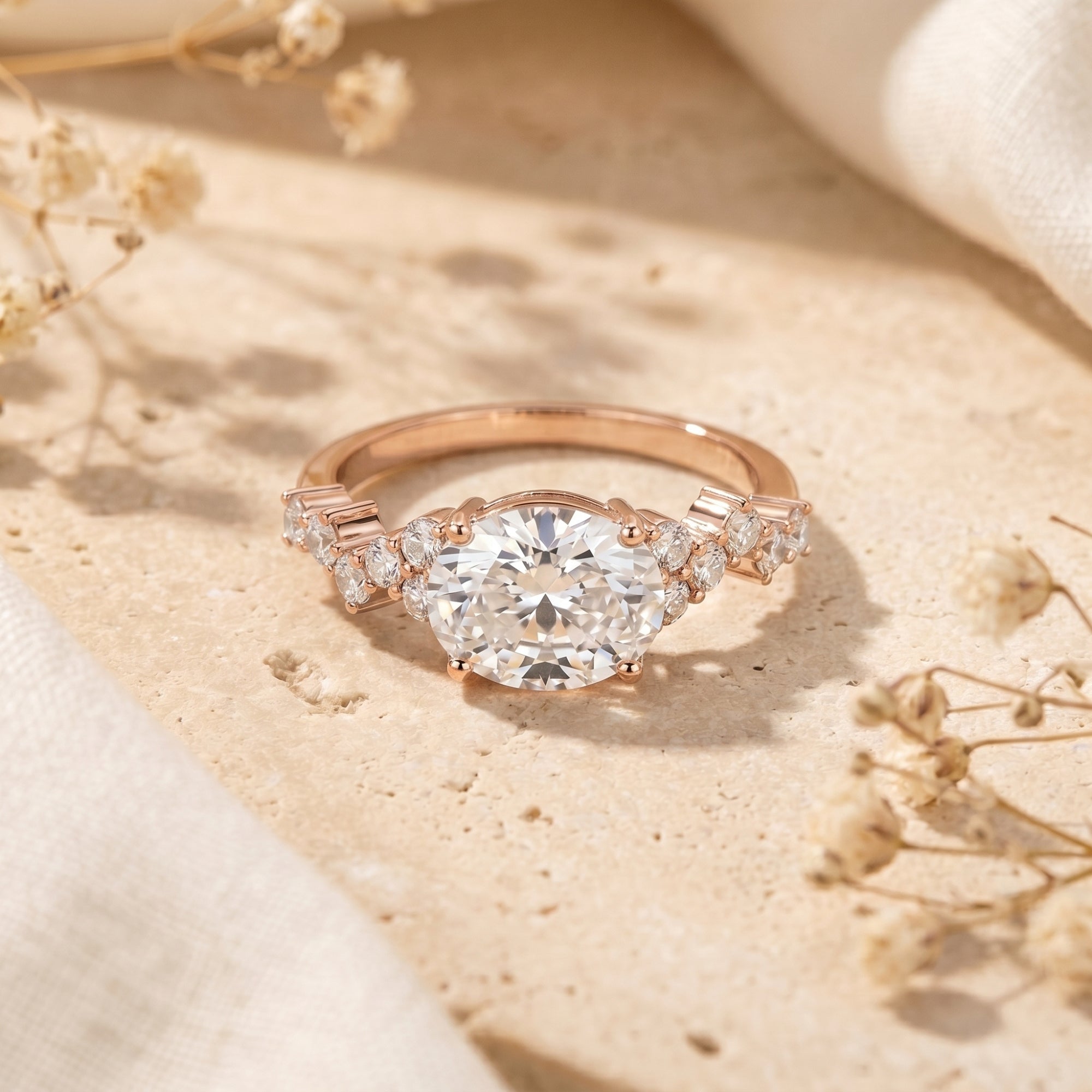 Rose gold oval moissanite promise ring with cluster pavé side stones on a stone surface with dried flowers.