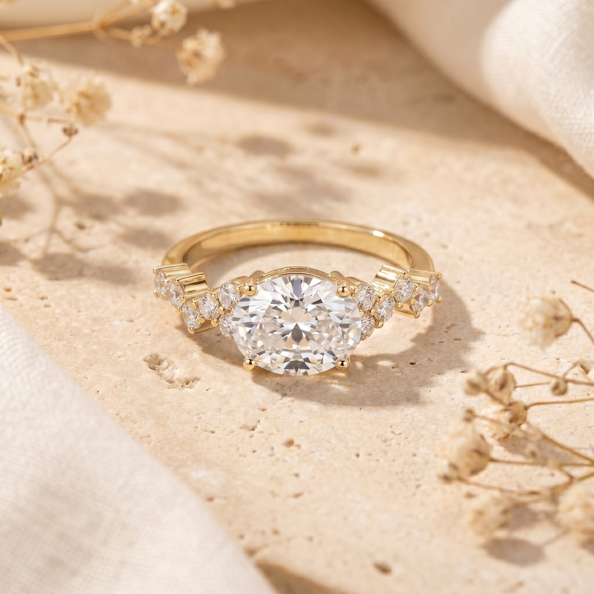 Yellow gold oval moissanite promise ring with cluster pavé side stones on a stone surface with dried flowers.