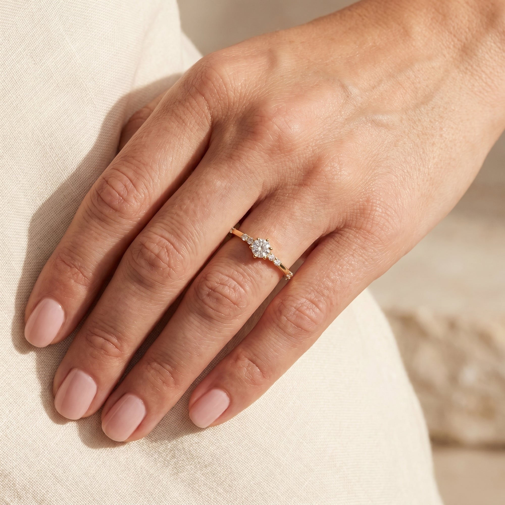 Model wearing a gold round cut moissanite engagement ring against a light linen fabric background.