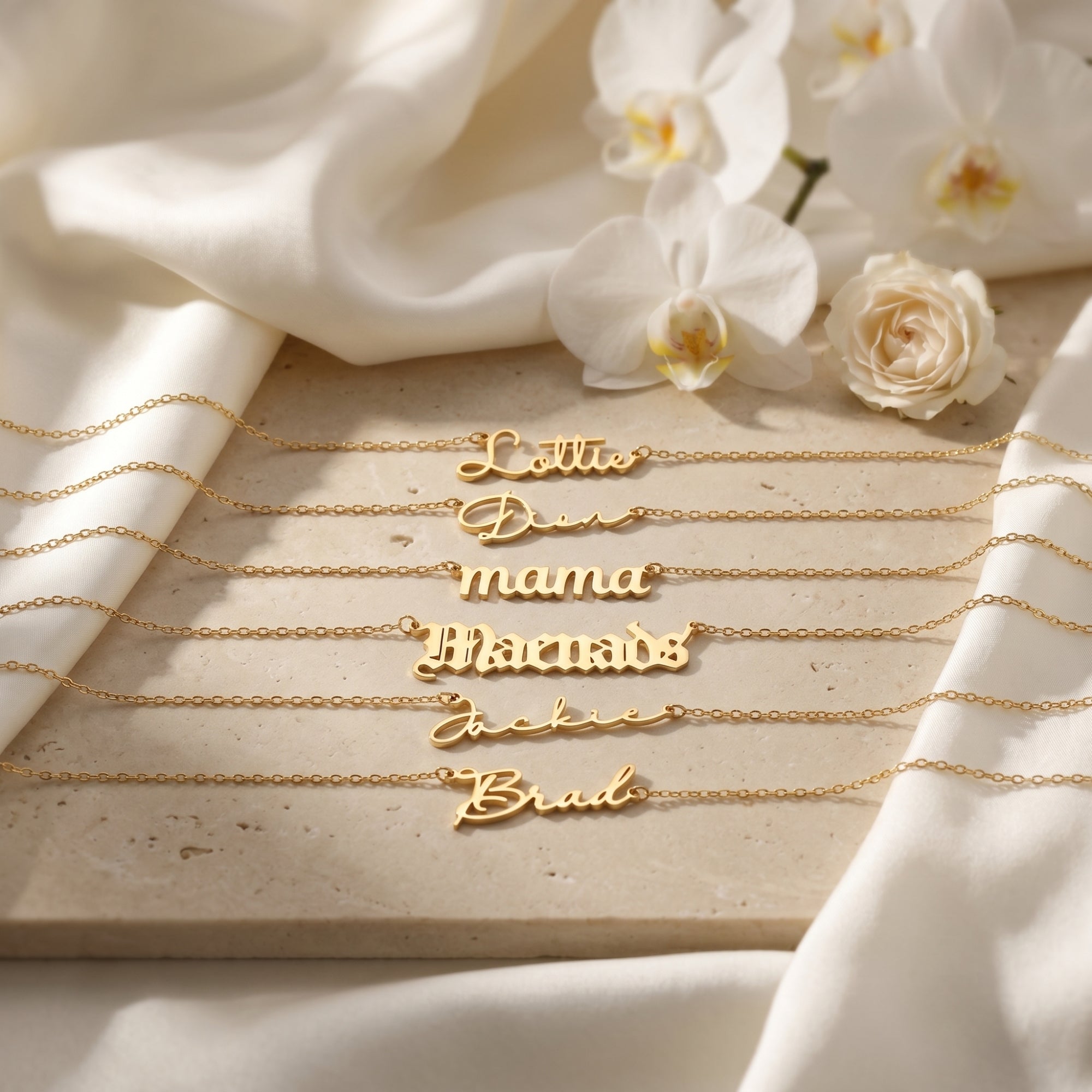 Six 18K gold finish personalized name necklaces with different font styles on a stone slab with white flowers.
