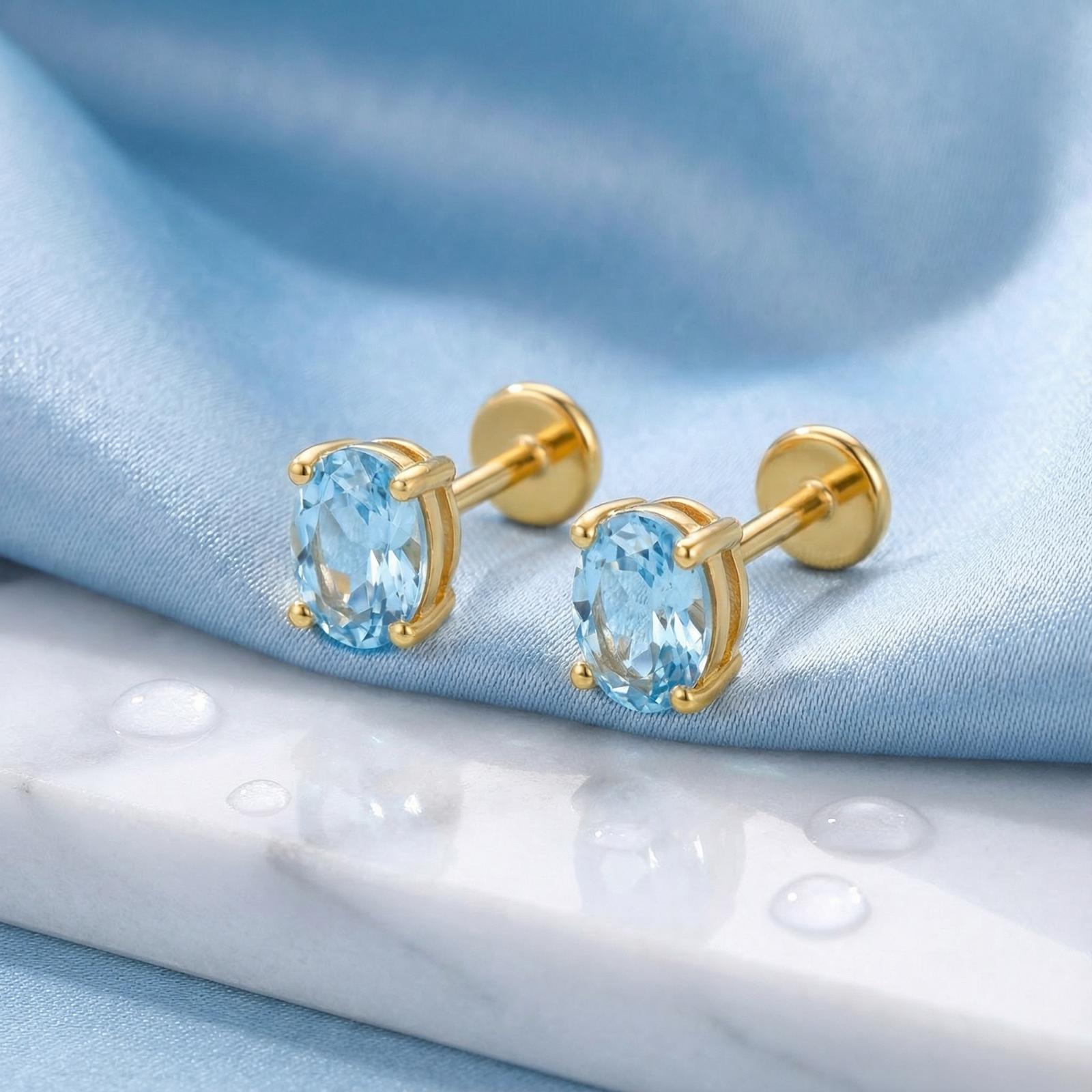 Side view of oval cut simulated aquamarine stud earrings in 18K gold vermeil on marble and blue silk.