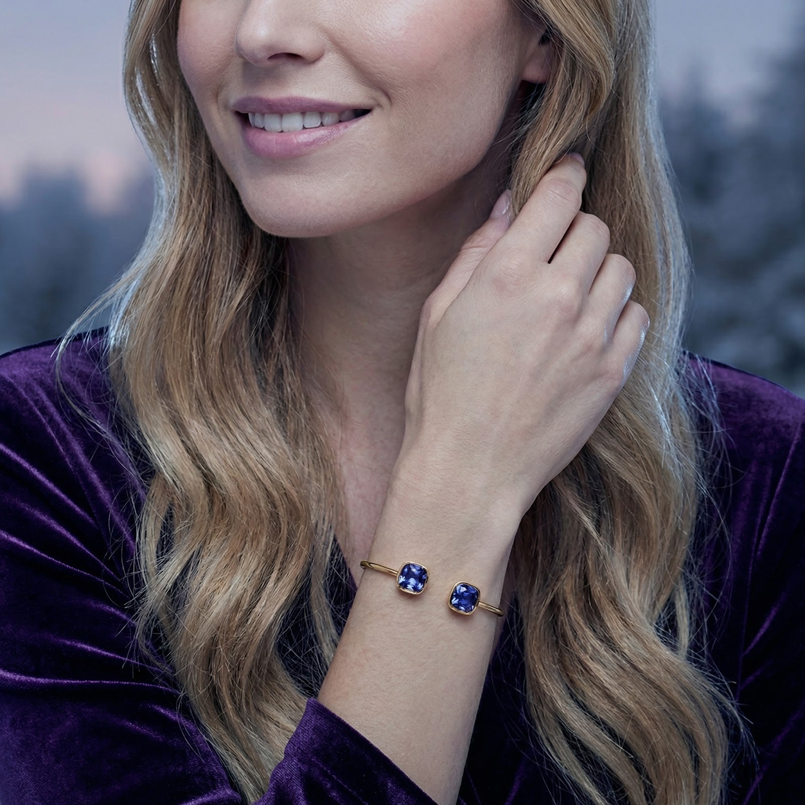 Gold Nerina bangle with blue simulated tanzanite stones worn on a wrist with a purple velvet sleeve.