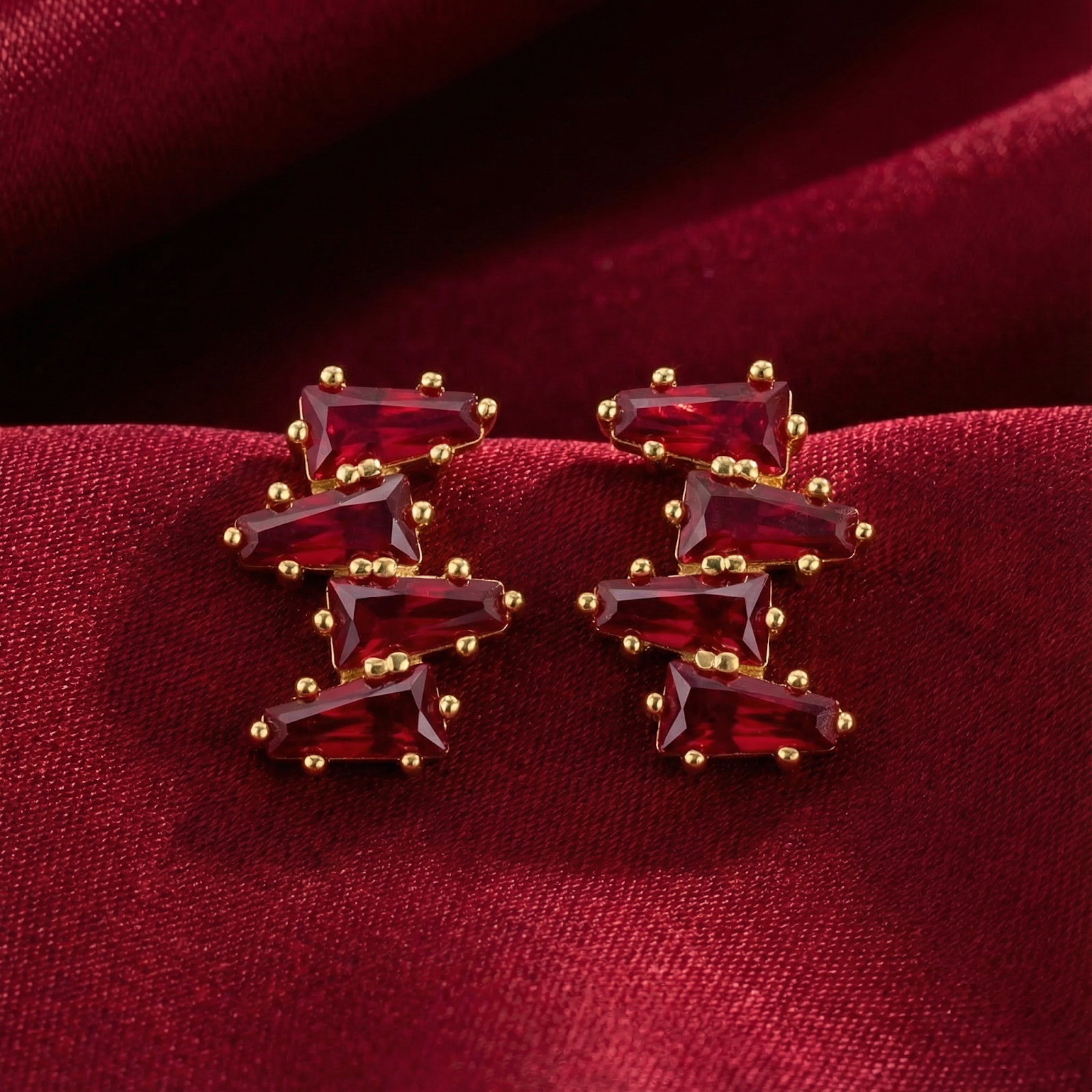 Front view of Rhianna Studs in 18K gold vermeil with red trillion-cut rubies on a red fabric background.