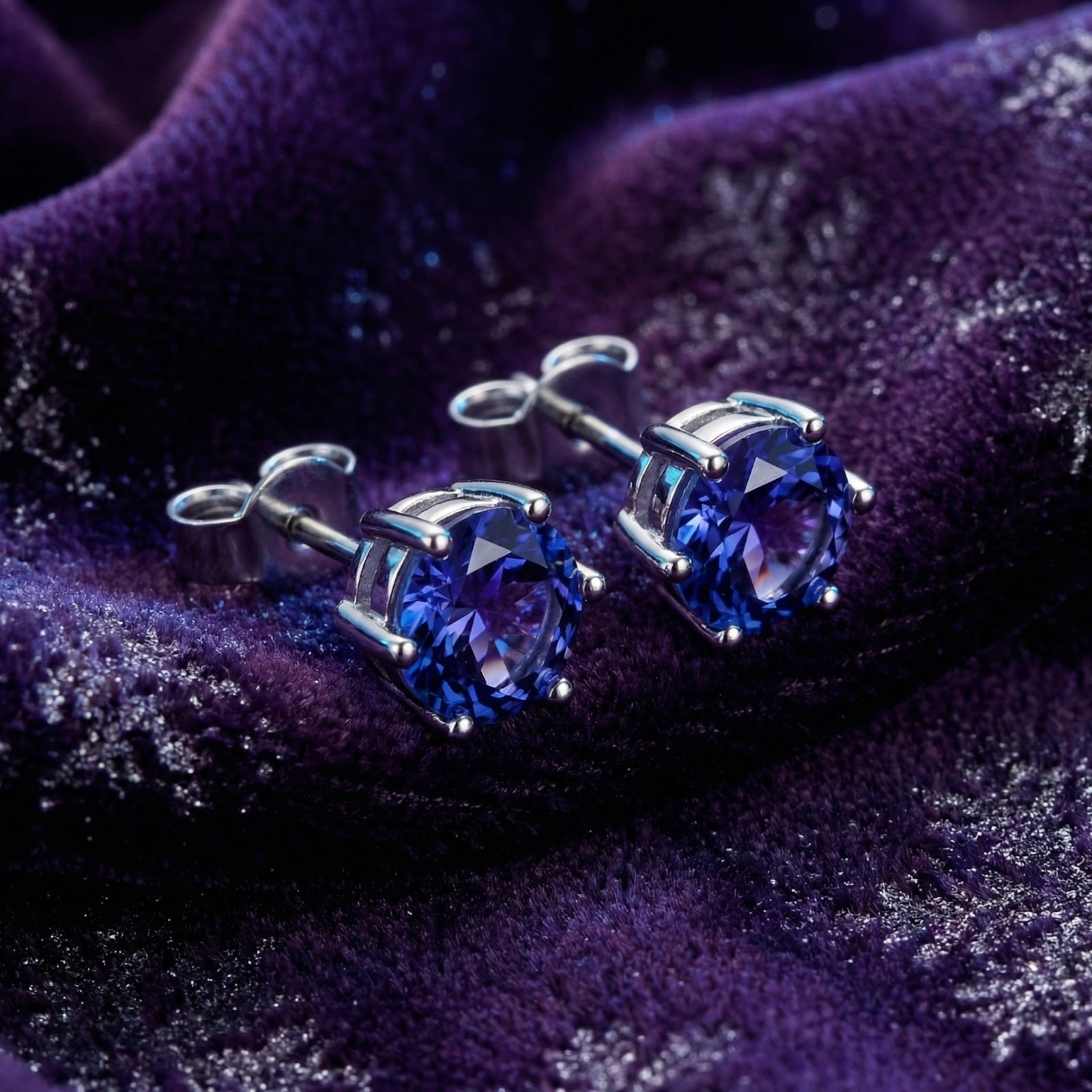 Round cut simulated tanzanite stud earrings in S925 sterling silver resting on purple velvet fabric.