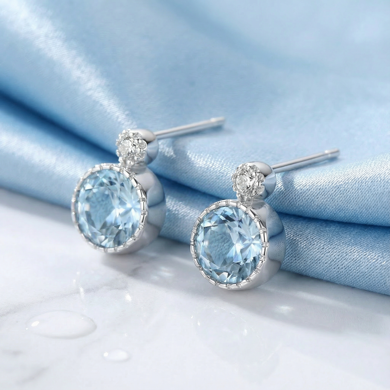 Angled view of round-cut simulated aquamarine stud earrings in sterling silver on blue silk and marble.
