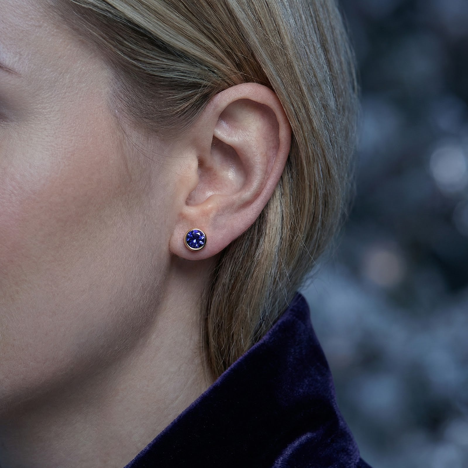 Model wearing a round blue simulated tanzanite stud earring in an 18K gold vermeil bezel setting.