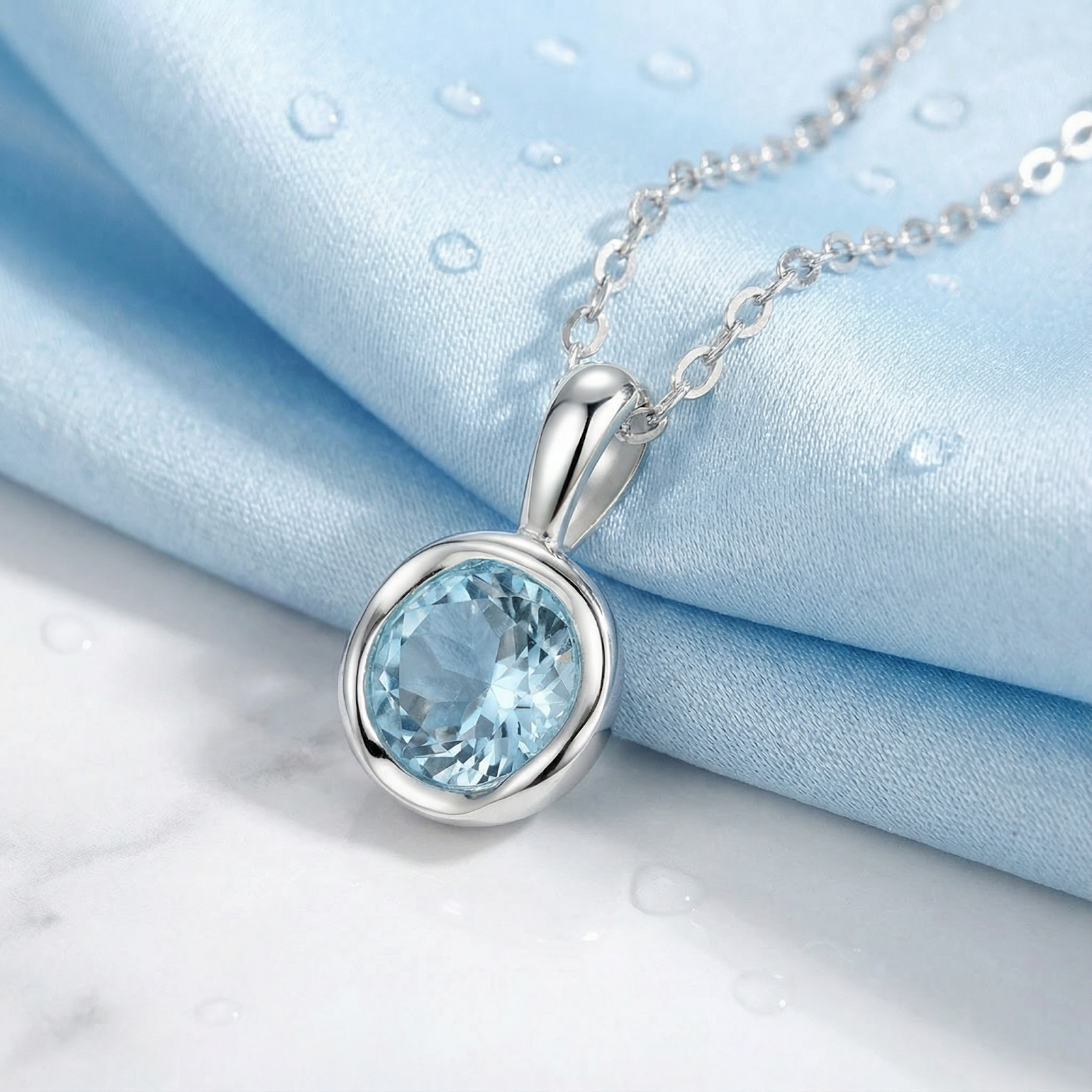 The Marella round-cut simulated aquamarine pendant in sterling silver on a light blue silk background.