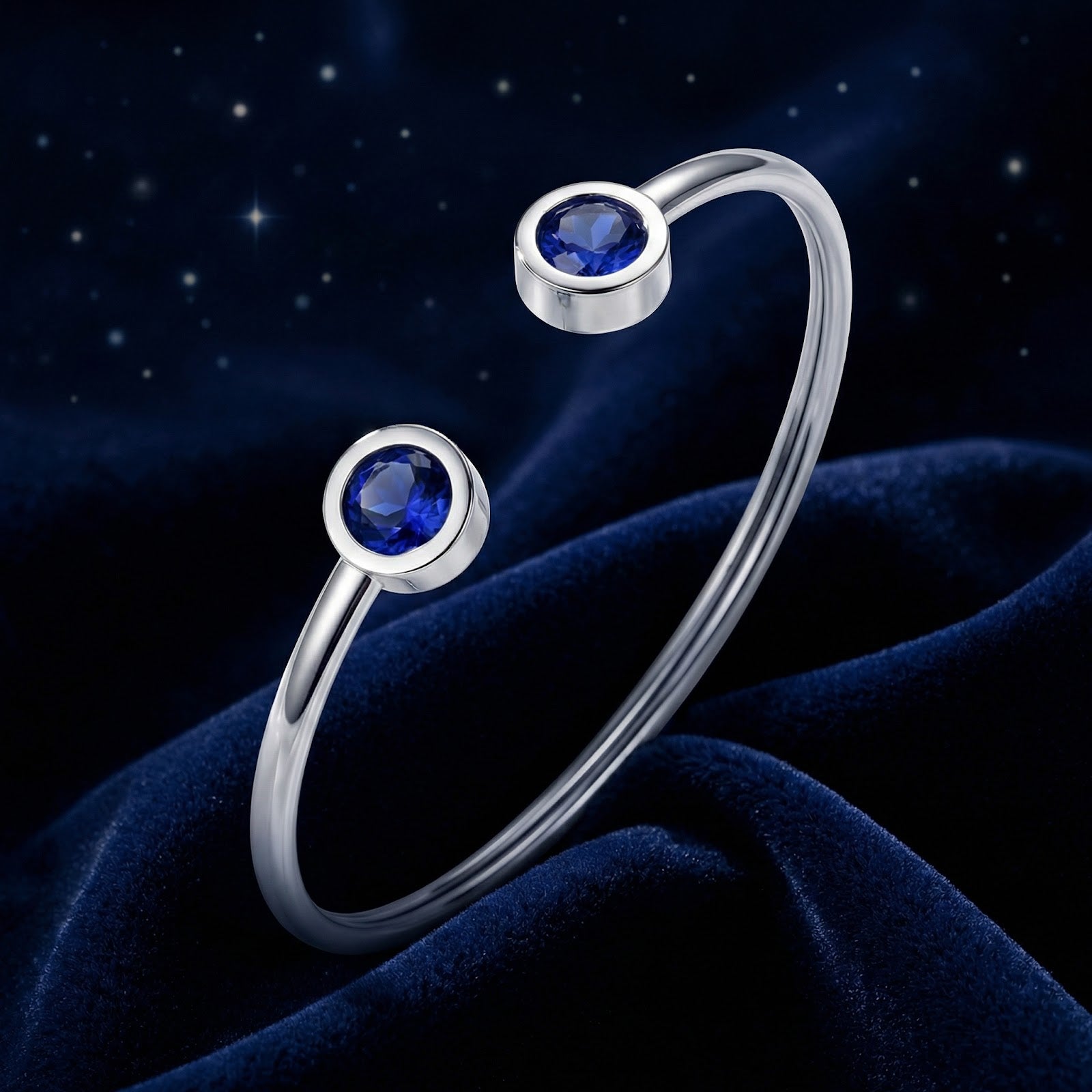 Silver stainless steel Verian bangle with two round blue simulated sapphires on a dark blue velvet background.