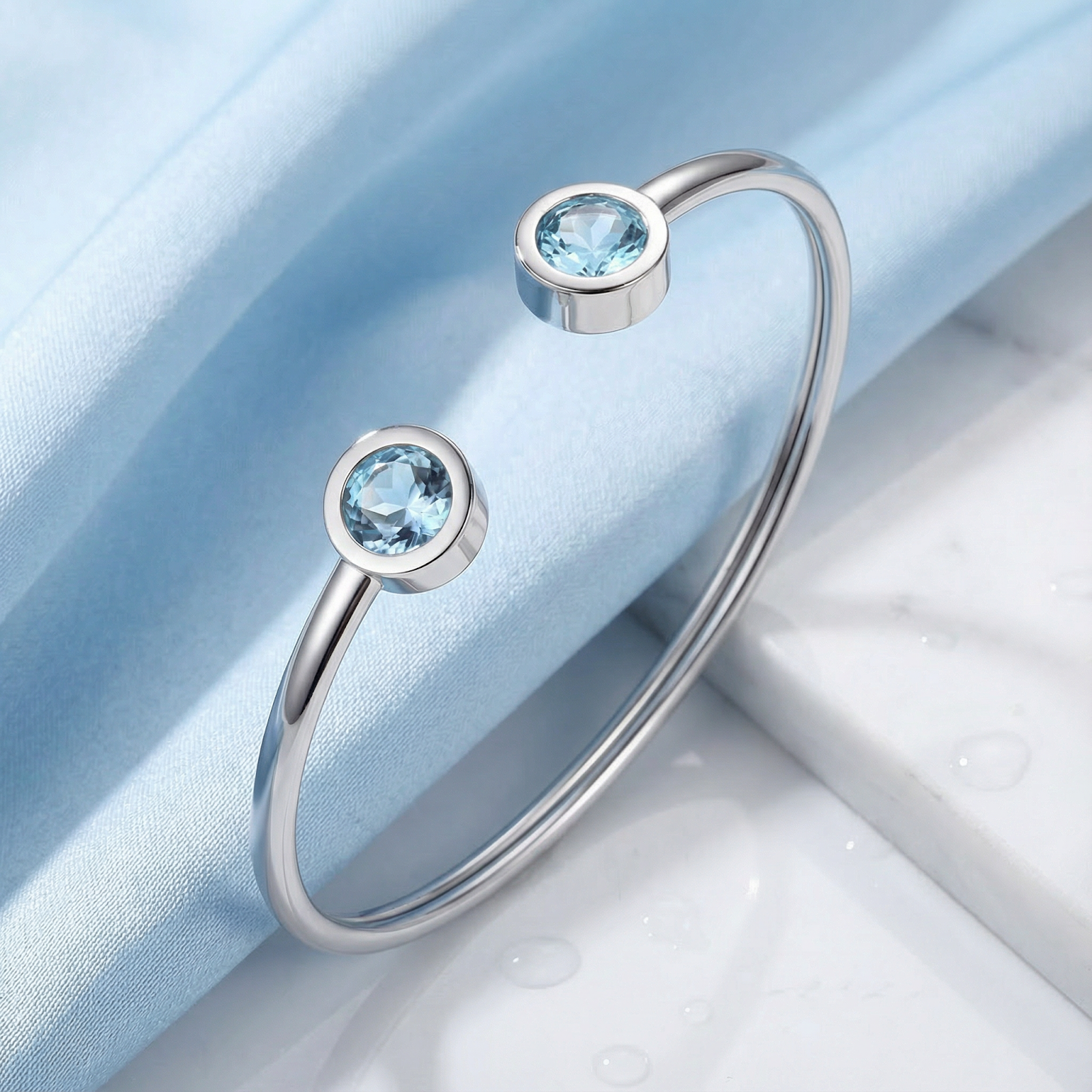 Silver stainless steel Cyrene bangle with round-cut simulated aquamarine stones on blue fabric and marble.