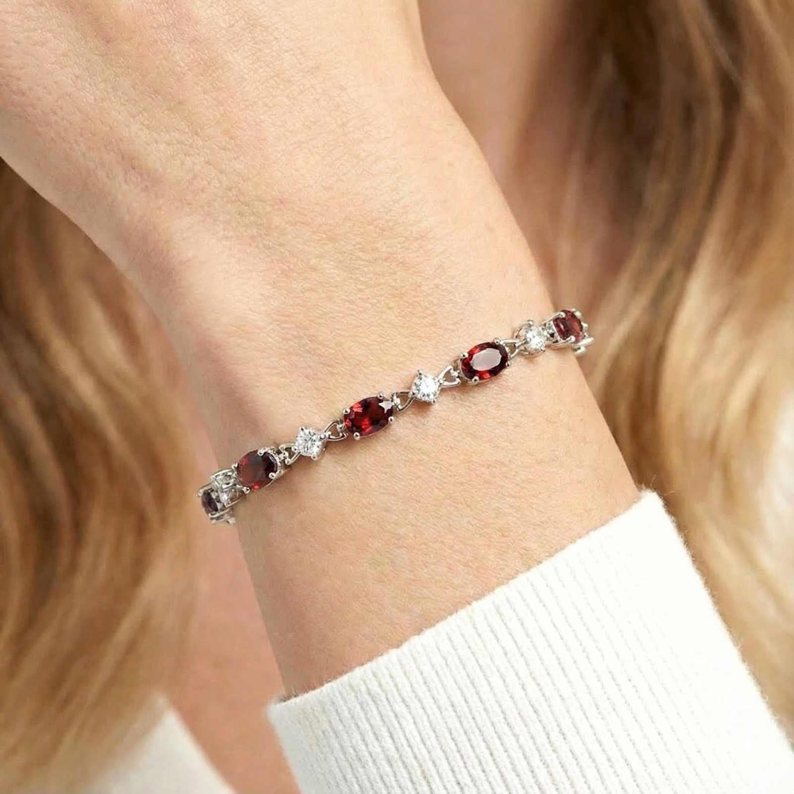 Close-up of oval-cut simulated garnets and round white stones on a silver station bracelet worn on a wrist.