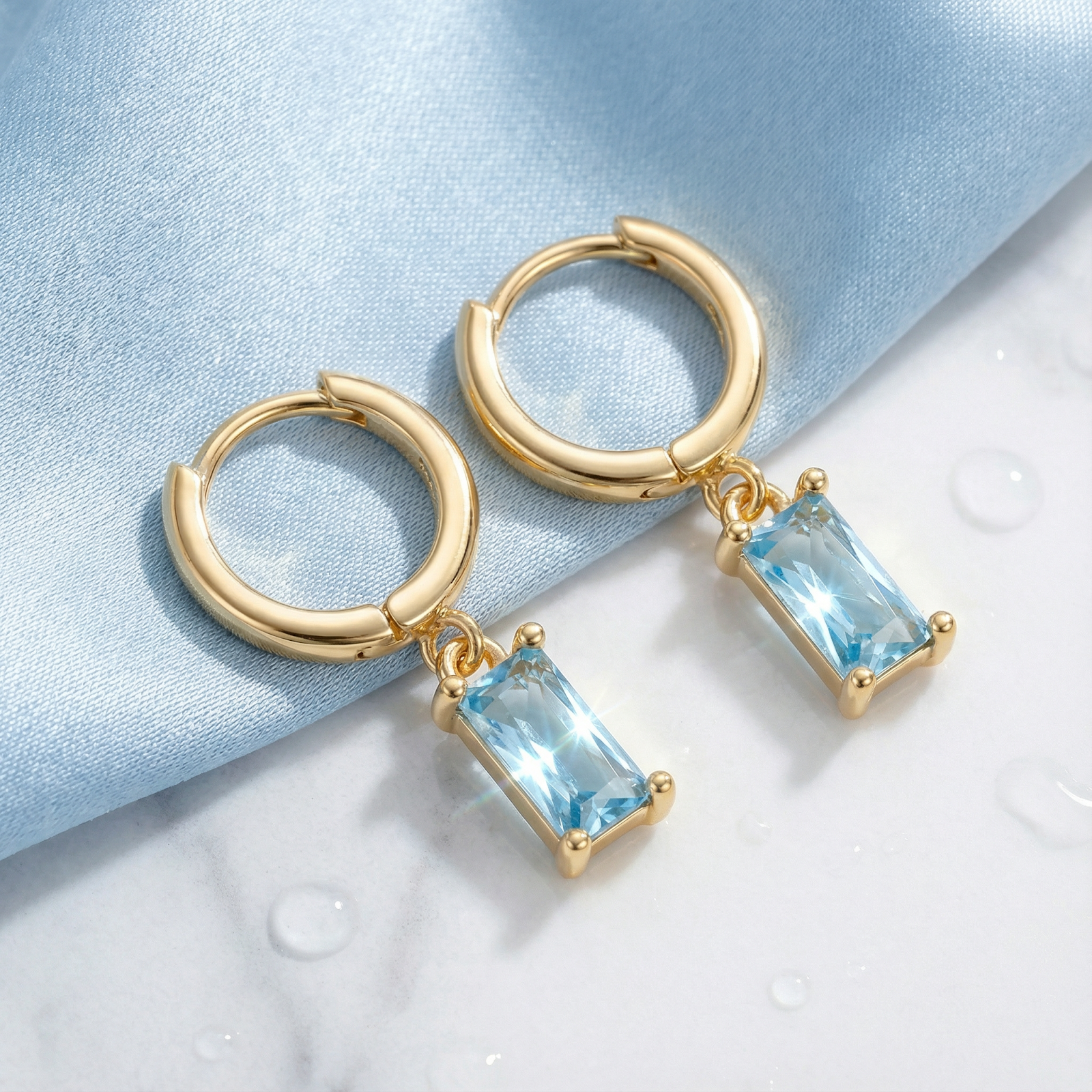 18K gold vermeil hoop earrings with rectangular-cut simulated aquamarine stones on blue fabric with water droplets.