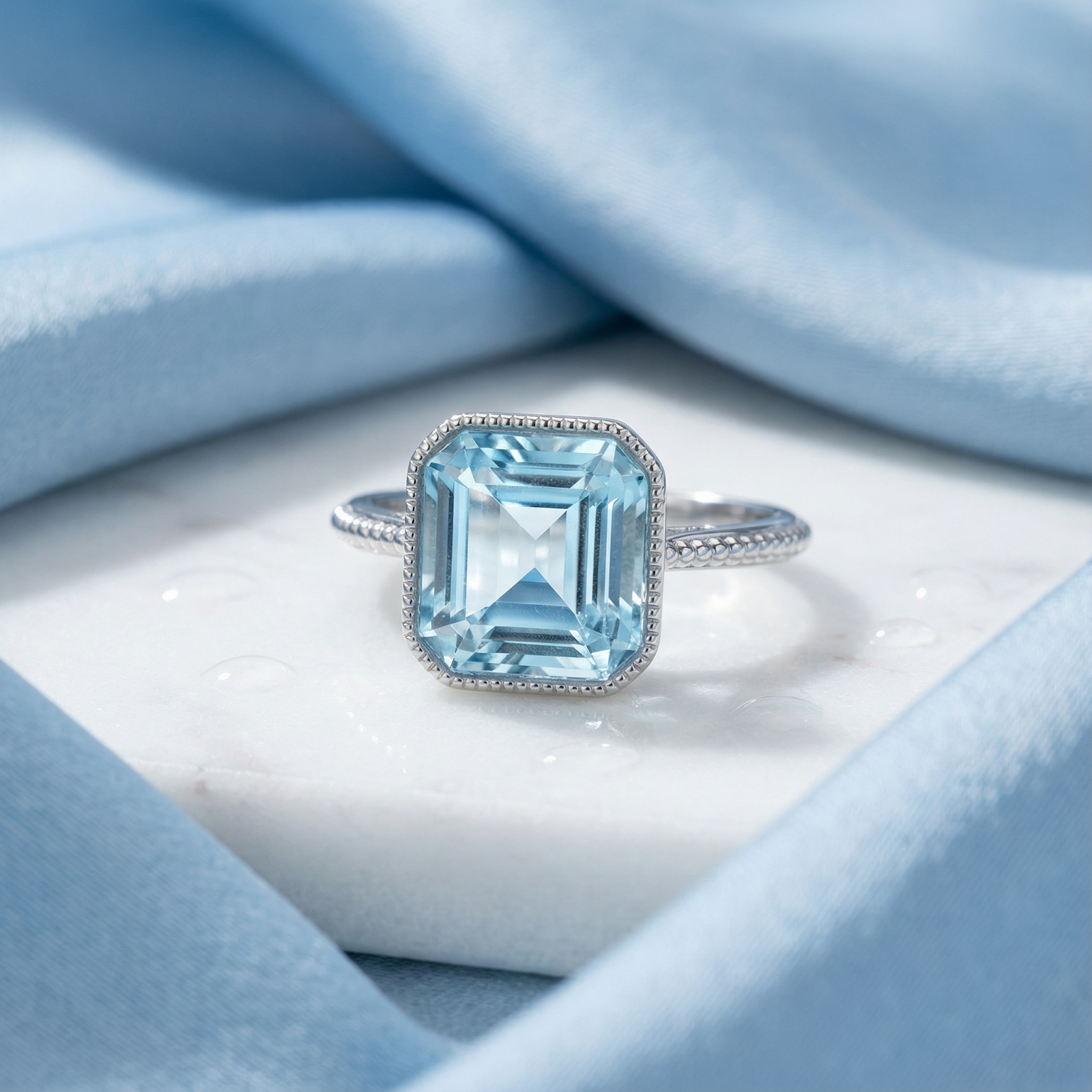 Close-up of a rectangular cut simulated aquamarine ring with a textured bezel setting on marble.