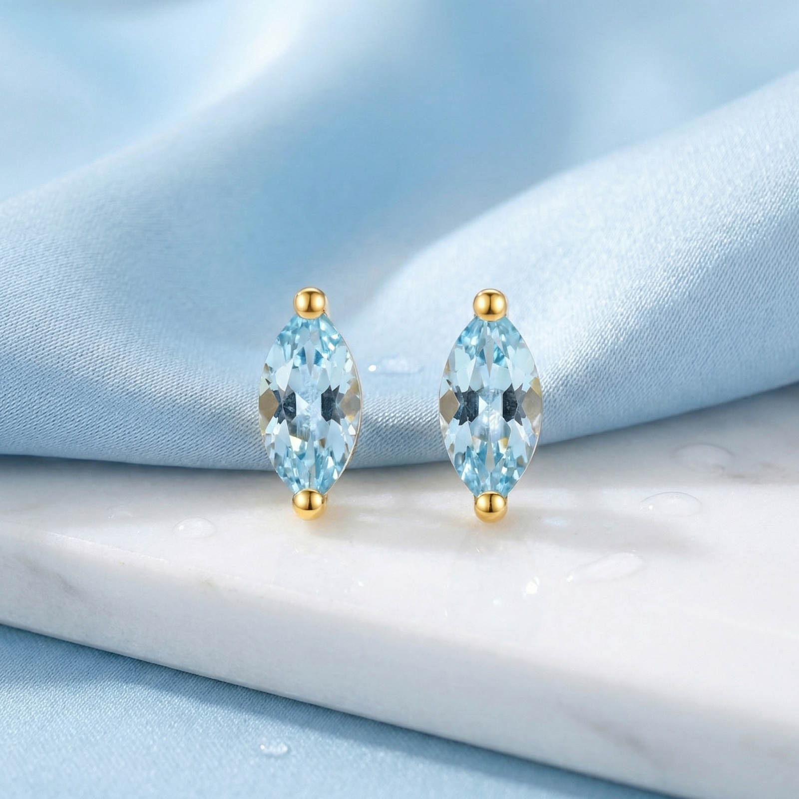 Front view of marquise cut simulated aquamarine earrings in 18K gold vermeil on marble and blue fabric.
