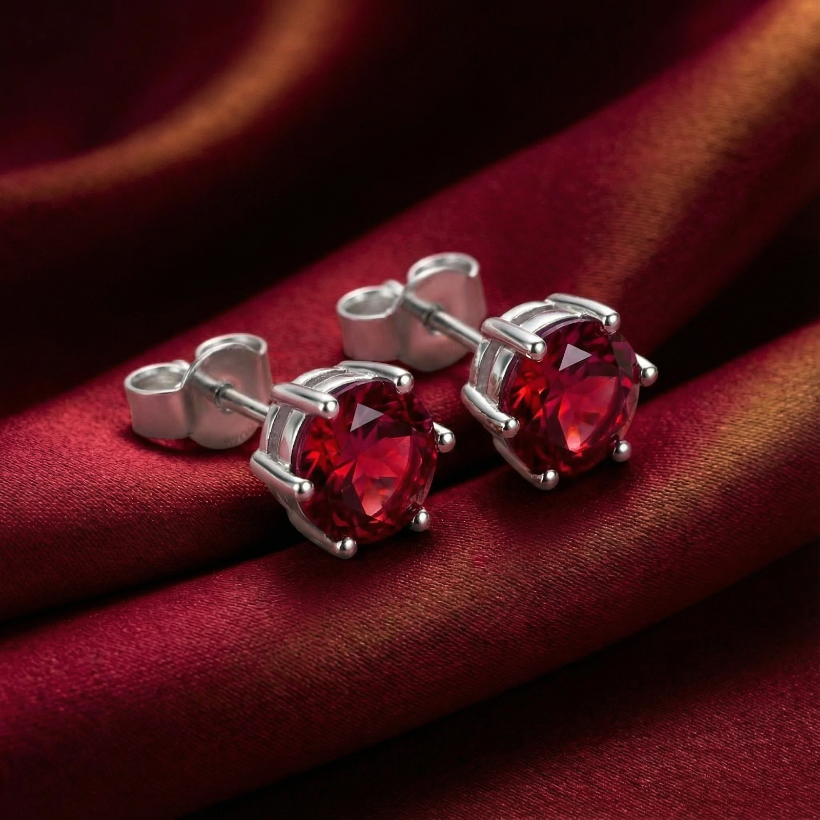 Round cut pigeon blood simulated ruby stud earrings in S925 sterling silver on red silk fabric.