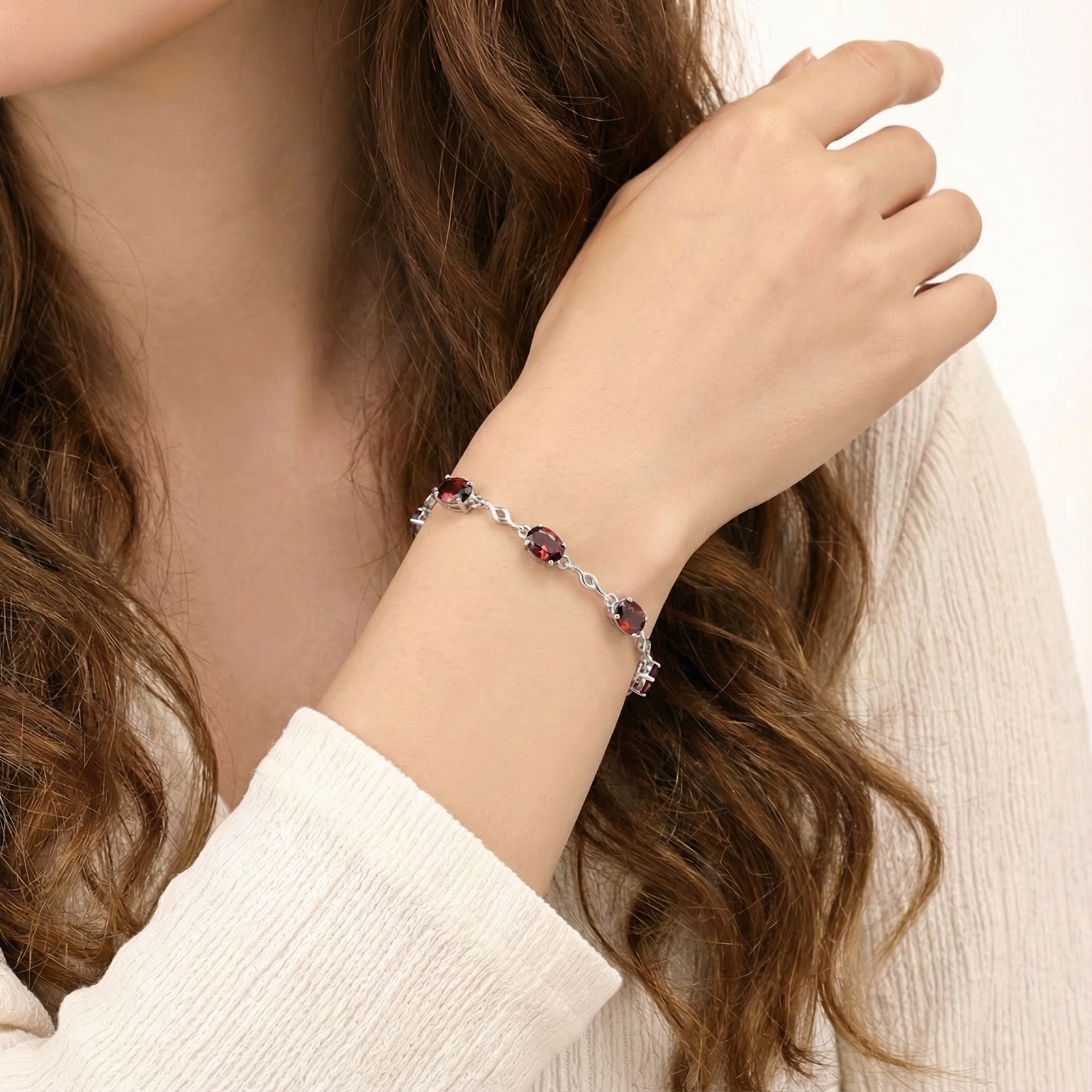 Close-up of the Valora silver station bracelet with oval-cut red garnets on a model's wrist wearing a cream sweater.
