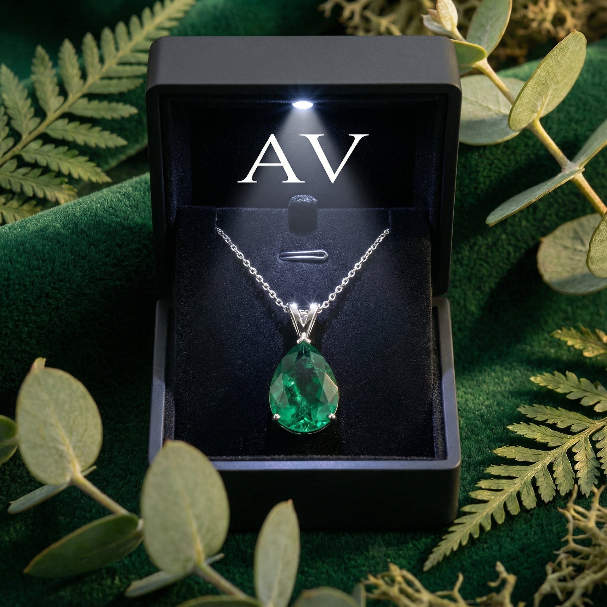 Green pear-cut moissanite necklace in a lighted black gift box on a bed of green velvet and leaves.