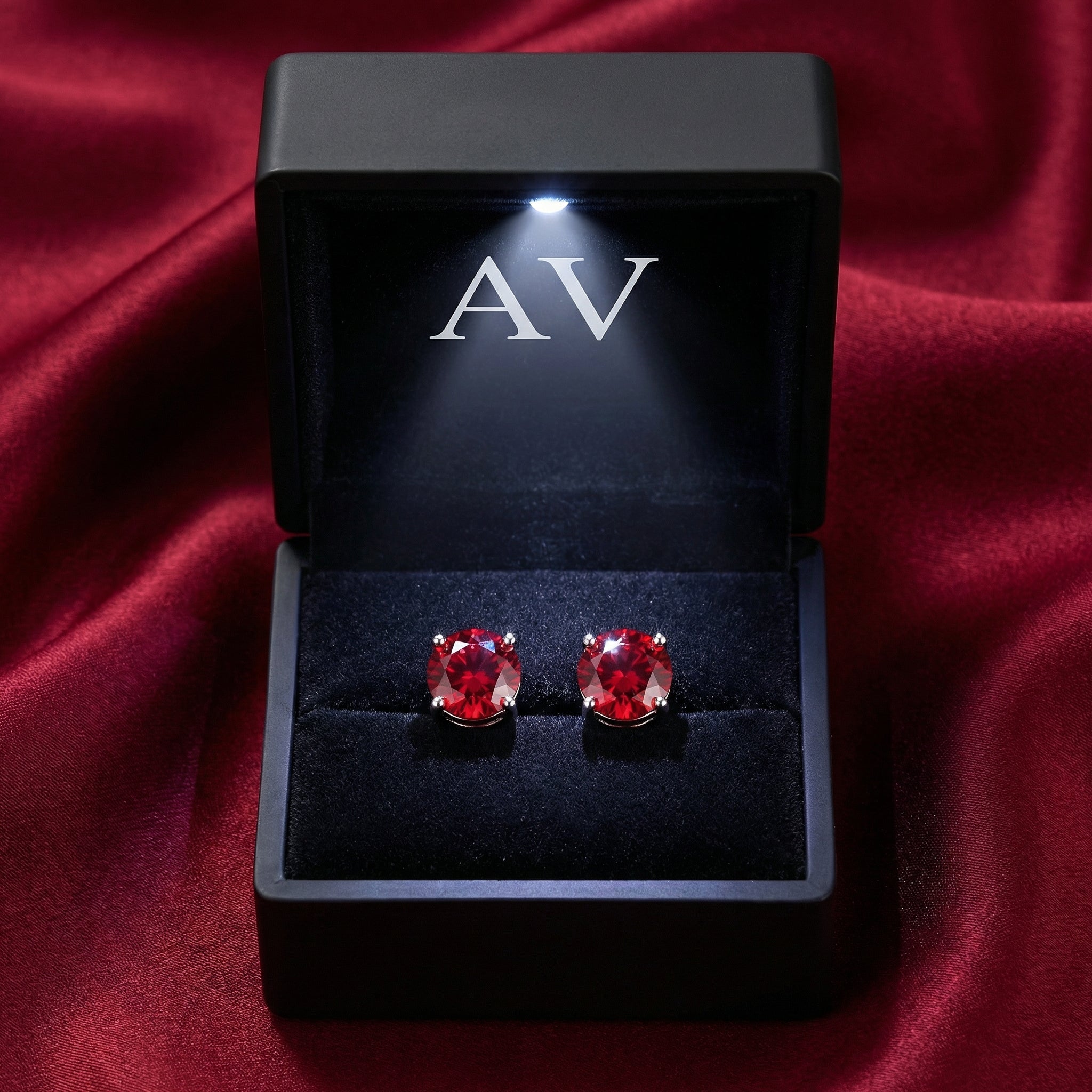 Round red simulated ruby stud earrings in a black LED-lit jewelry box on a red silk background.