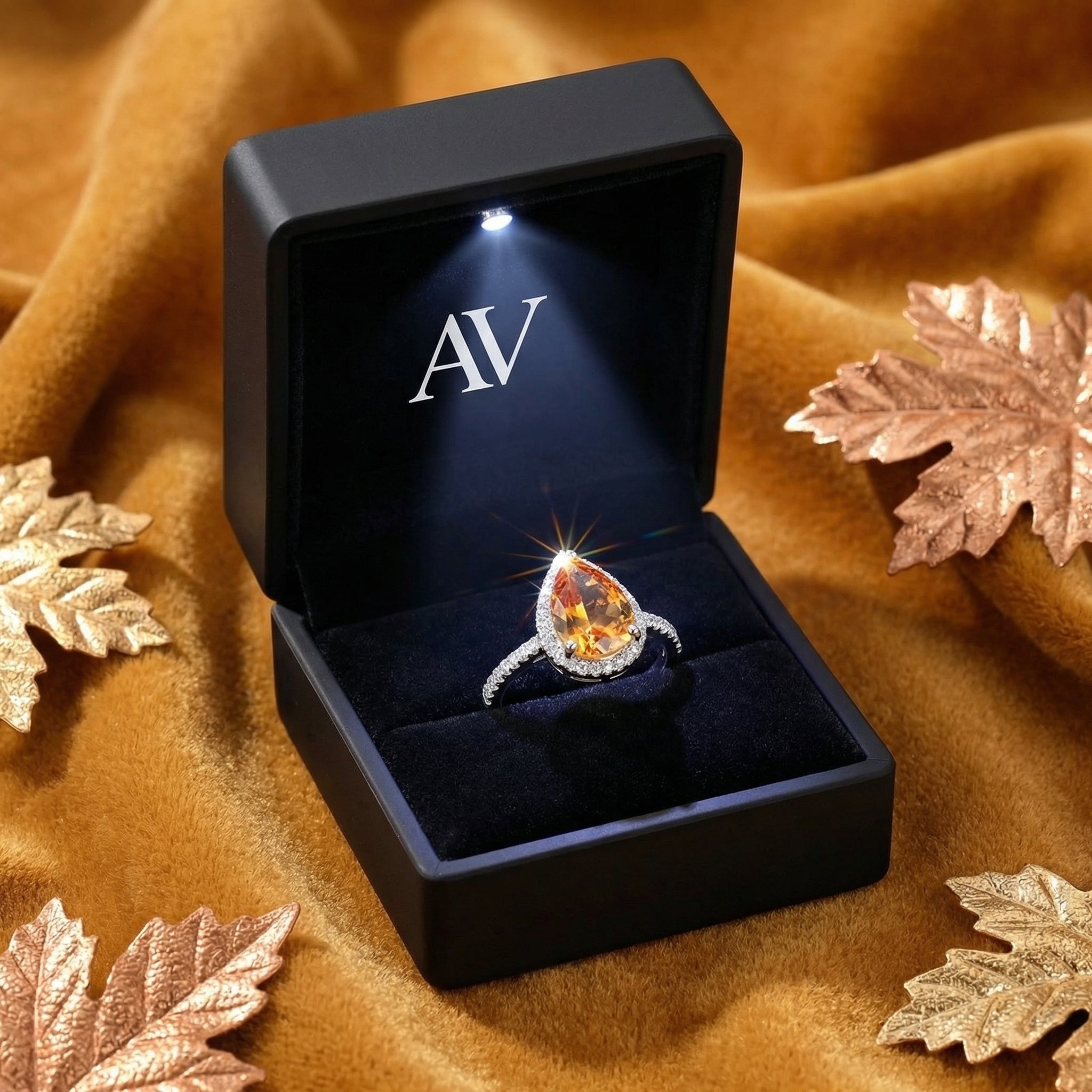Pear-cut simulated topaz ring in a black LED-lighted jewelry box with AV logo, set on a warm velvet background.