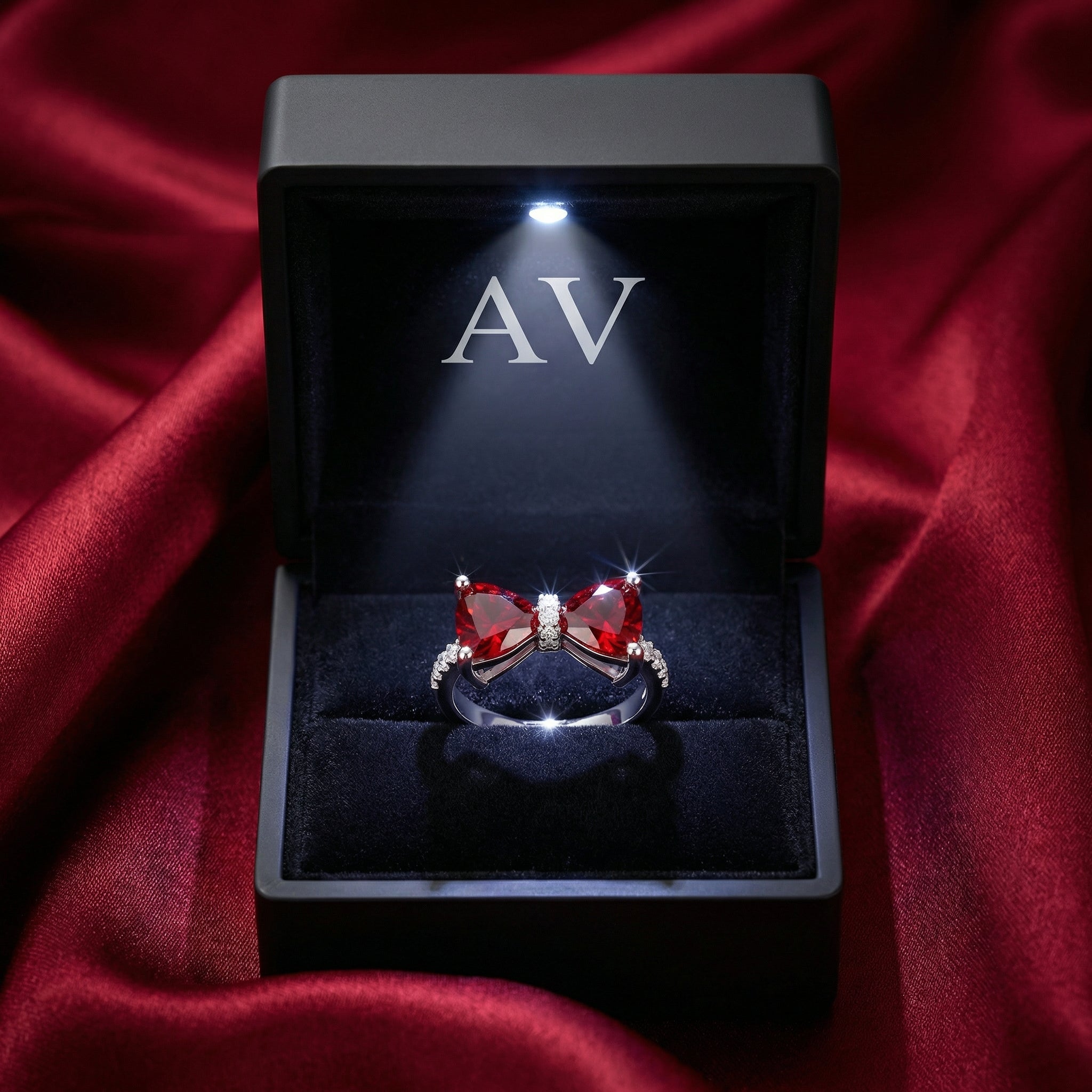 Vivara bow ring with red simulated rubies inside a lighted black AV jewelry box on red satin.