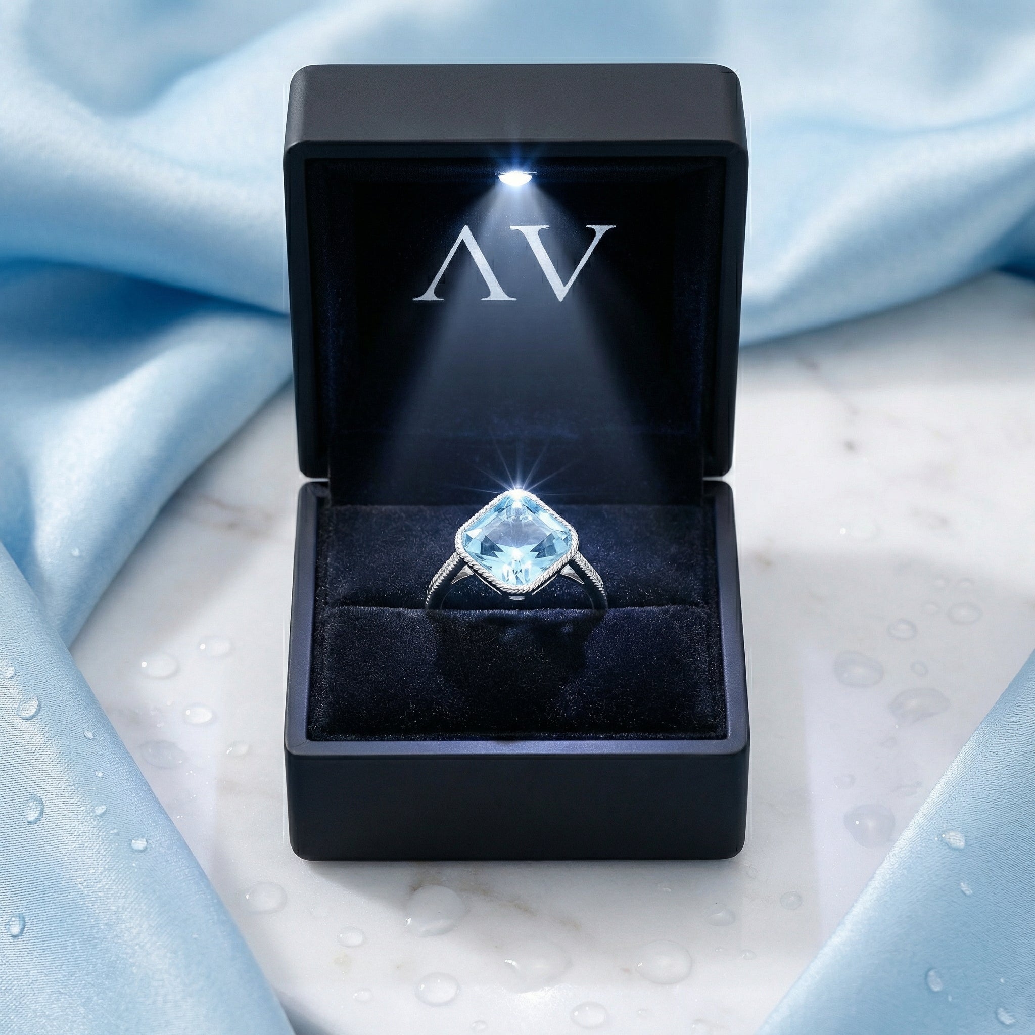 The Aquila cushion ring presented in a black LED-lighted jewelry box on a blue fabric background.