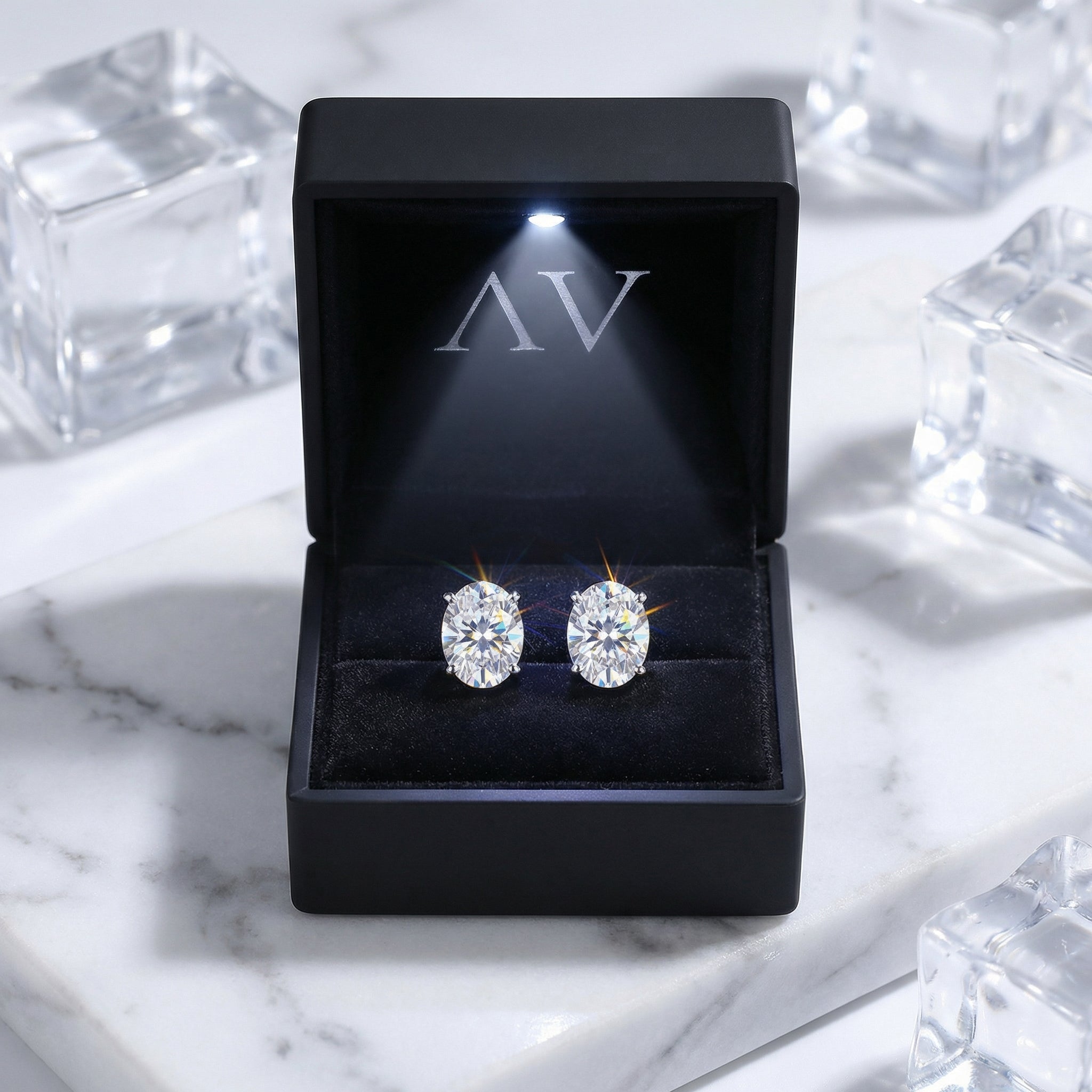 Oval cut zirconia stud earrings in a lighted black AV jewelry gift box on a white marble surface.