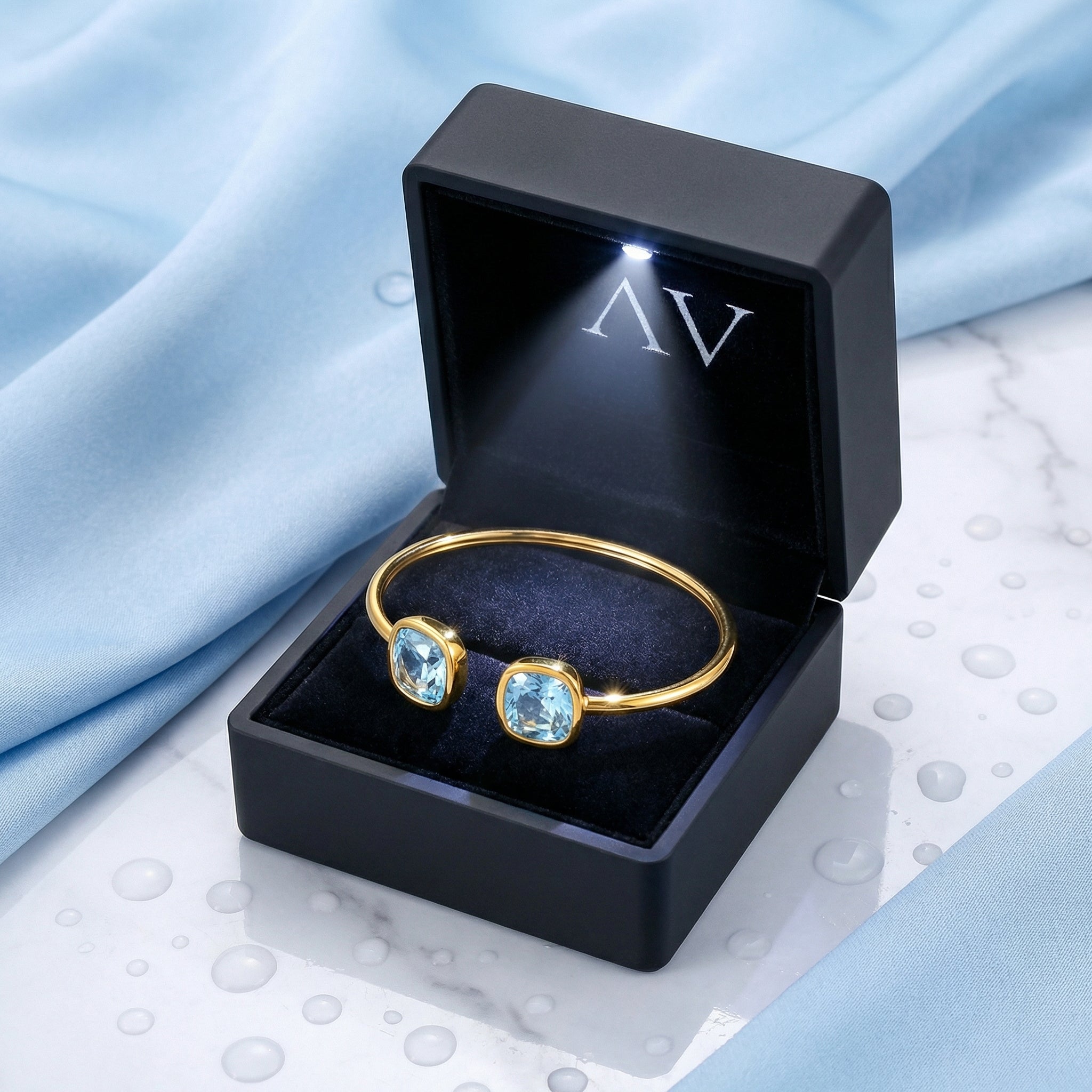 Gold bypass bangle with blue cushion-cut stones presented in a lighted black jewelry gift box.