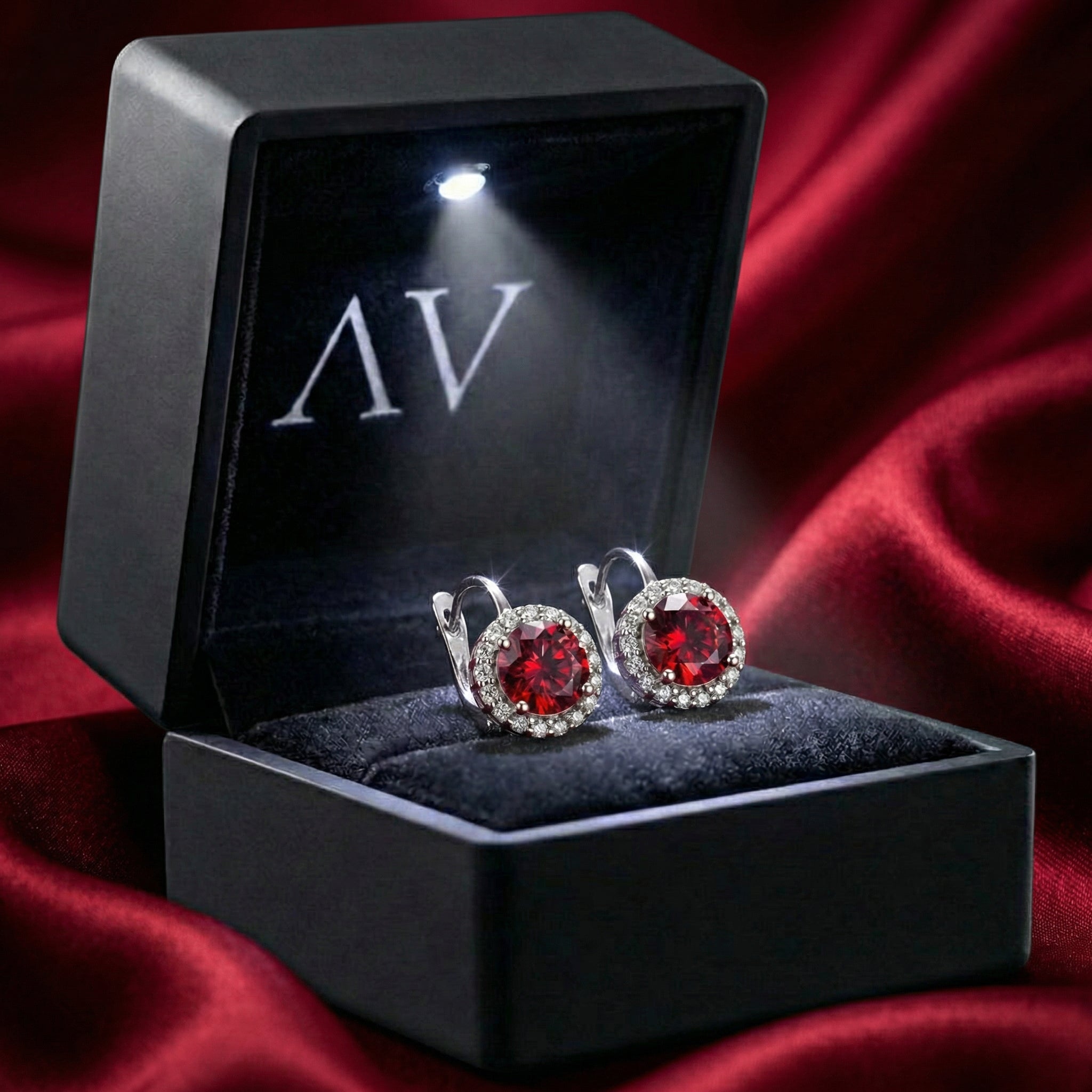 Pair of red ruby halo earrings in a black LED-lit gift box on a red satin background.