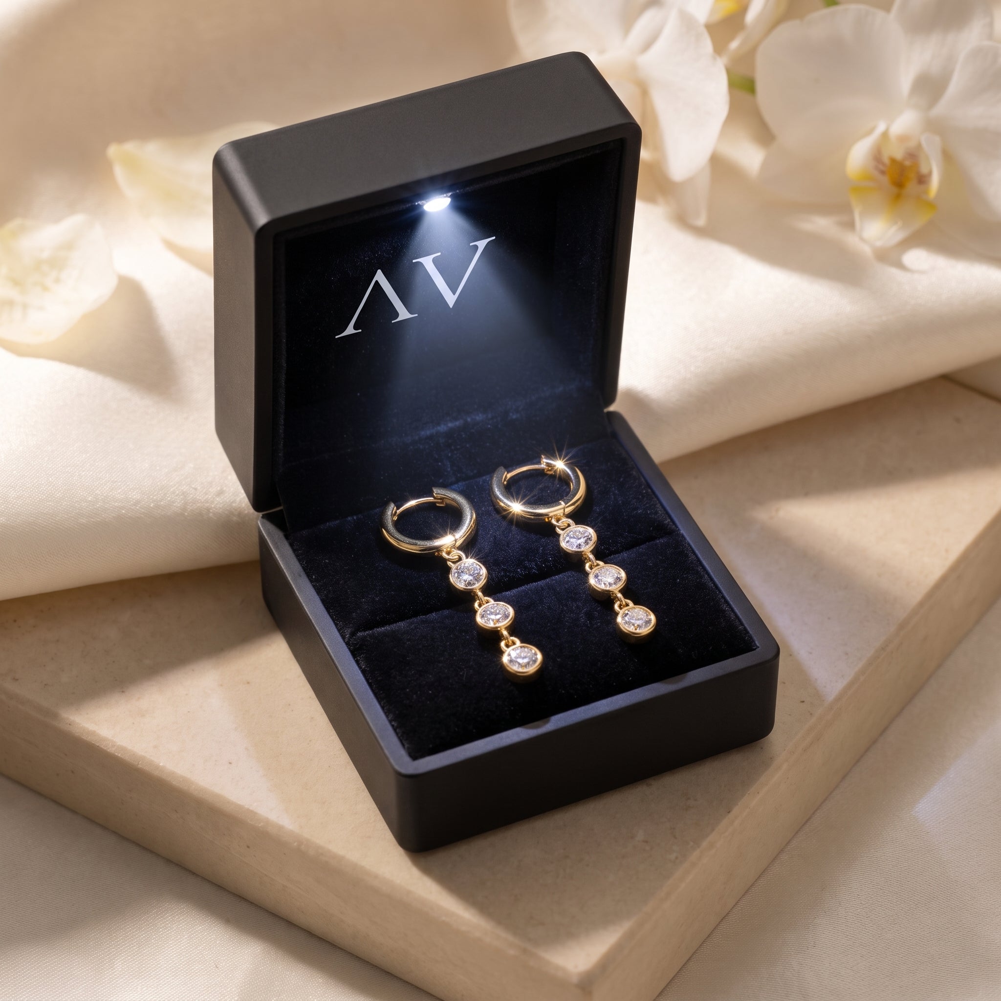 Gold moissanite drop earrings in a black LED-lit gift box on a cream fabric background.