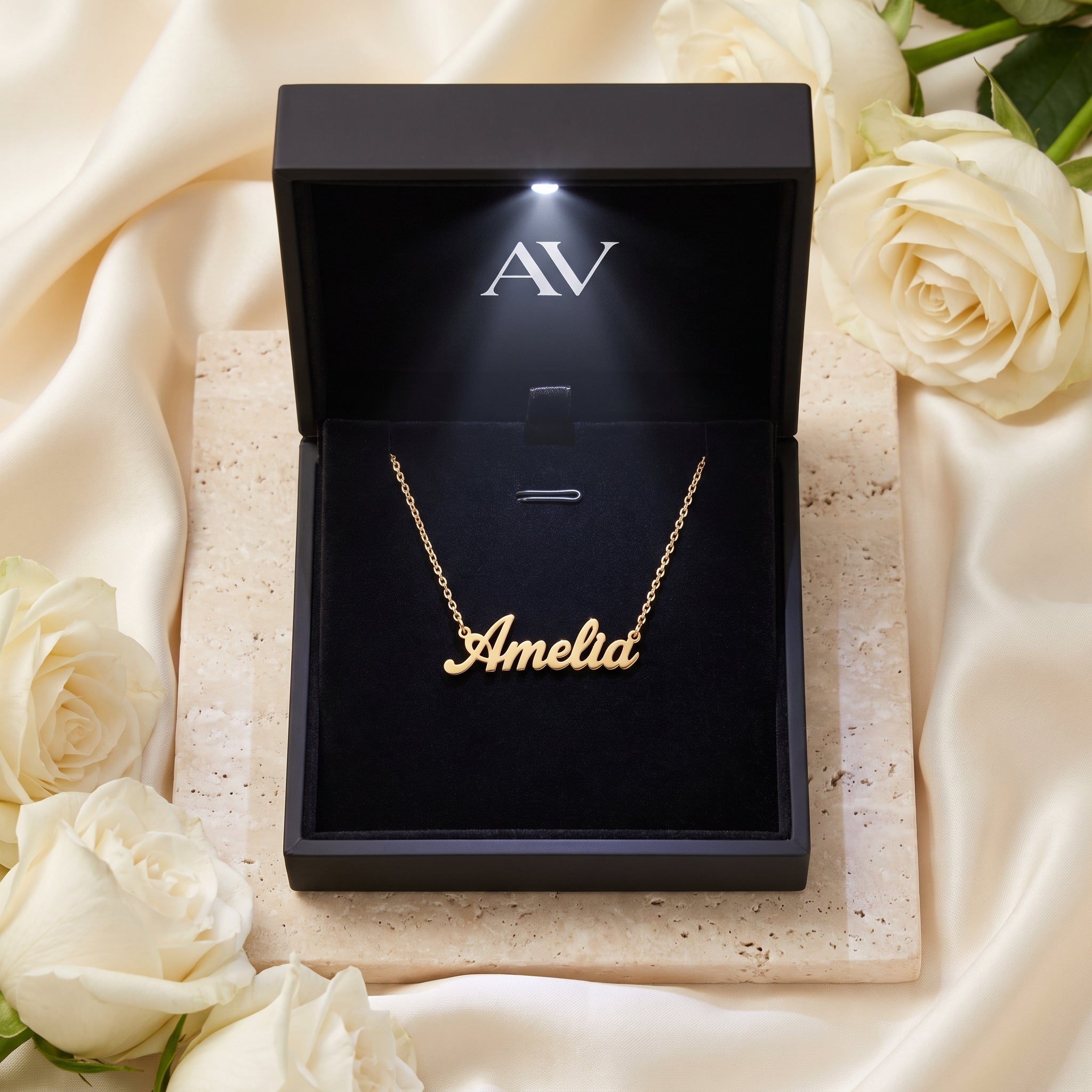 An 18K gold finish Amelia name necklace in a luxury black LED-lit gift box surrounded by white roses.