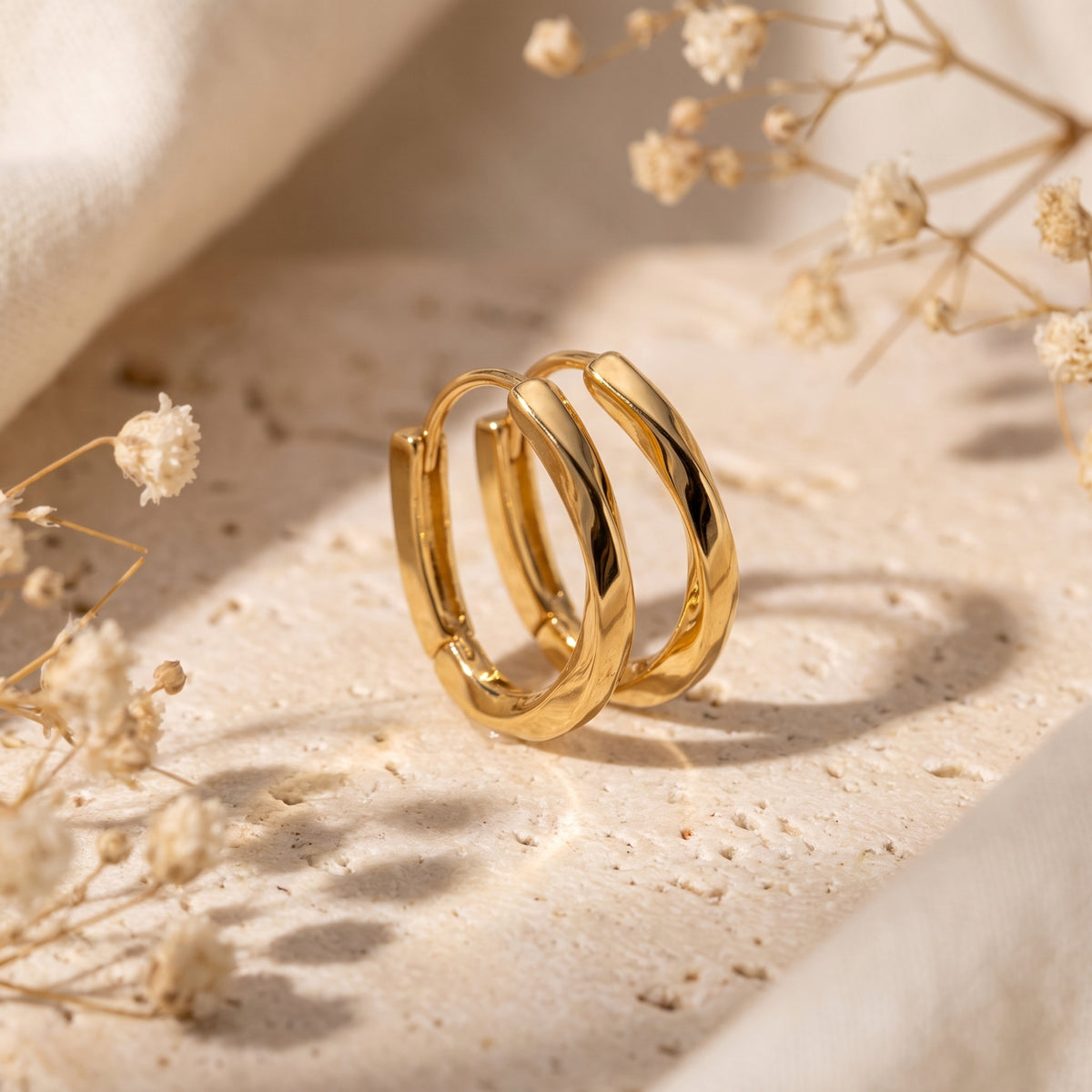 Gold-toned twisted hoop earrings standing upright together on a textured beige surface with dried flowers.