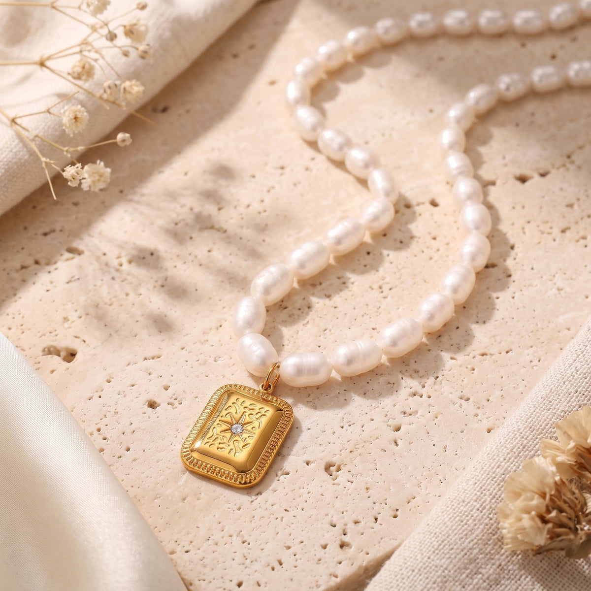 Freshwater pearl necklace with a rectangular gold-tone starburst pendant, displayed on a textured stone surface.