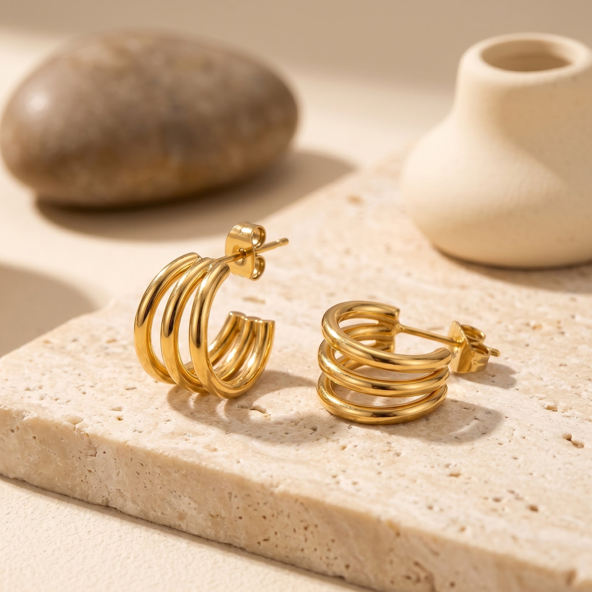 Gold triple-band hoop earrings on a stone slab with lifestyle props.