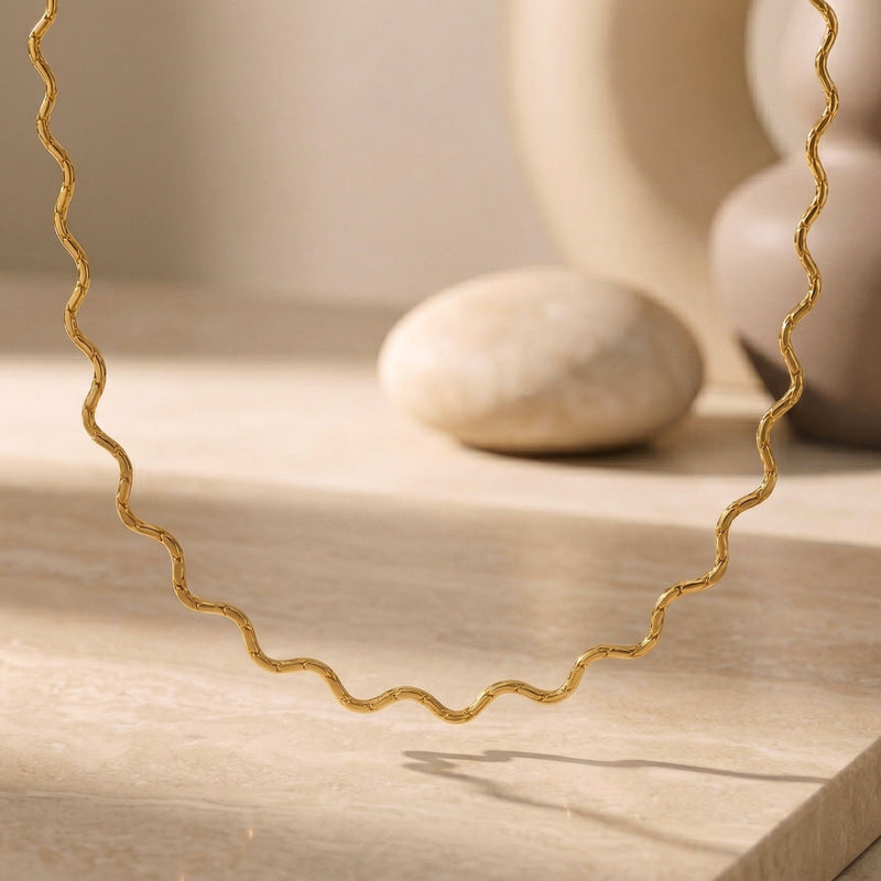 Gold-toned wavy chain necklace draped in a loop above a pale stone slab and rounded stone.