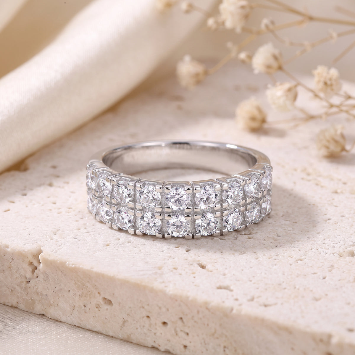 Double-row silver band ring set with two rows of round clear stones, resting on a beige stone slab.