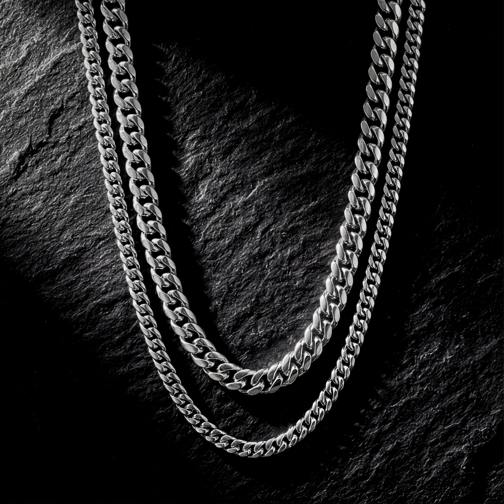 Two polished silver stainless steel Cuban link chains in a stacked set displayed on a dark textured stone background.