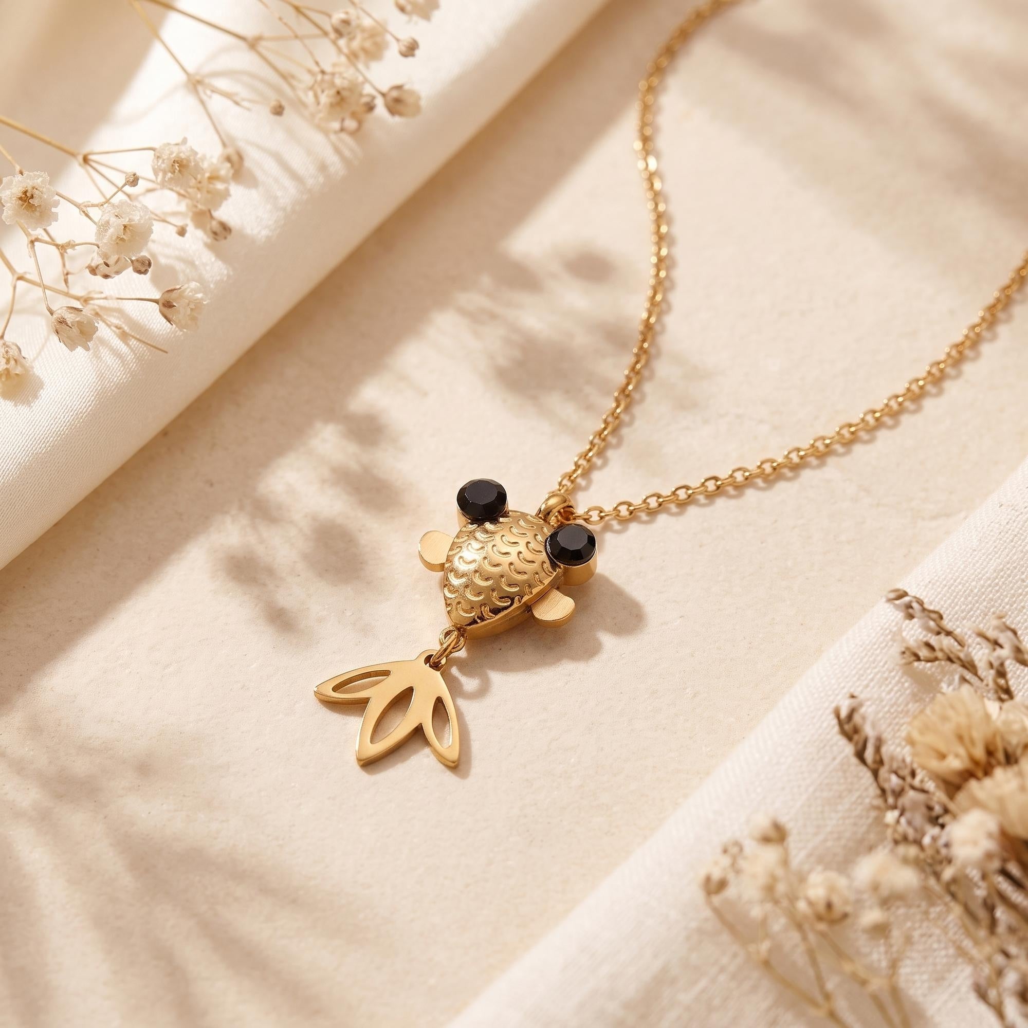 Gold finish stainless steel goldfish necklace with black eyes on a neutral fabric lifestyle background.