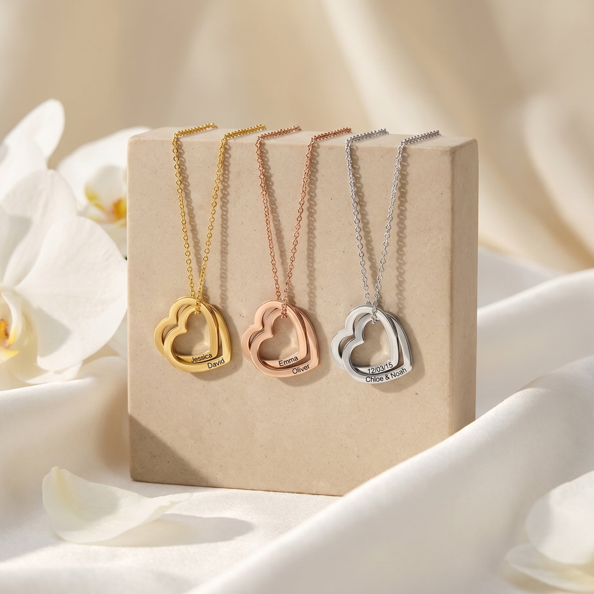 Gold, rose gold, and silver interlocking heart necklaces on a neutral block with white floral accents.
