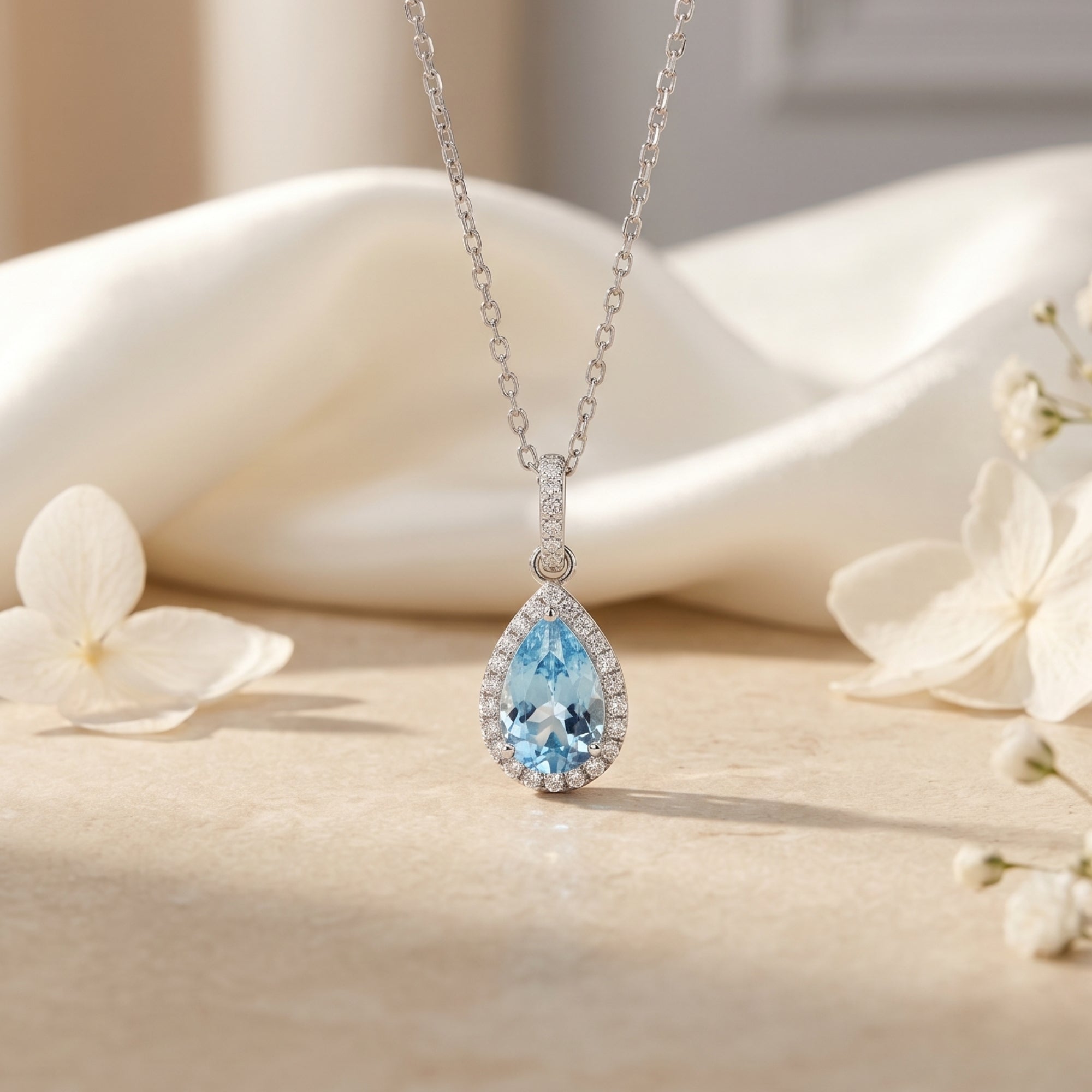 Pear-cut aquamarine halo necklace in sterling silver displayed on soft white fabric with flowers.