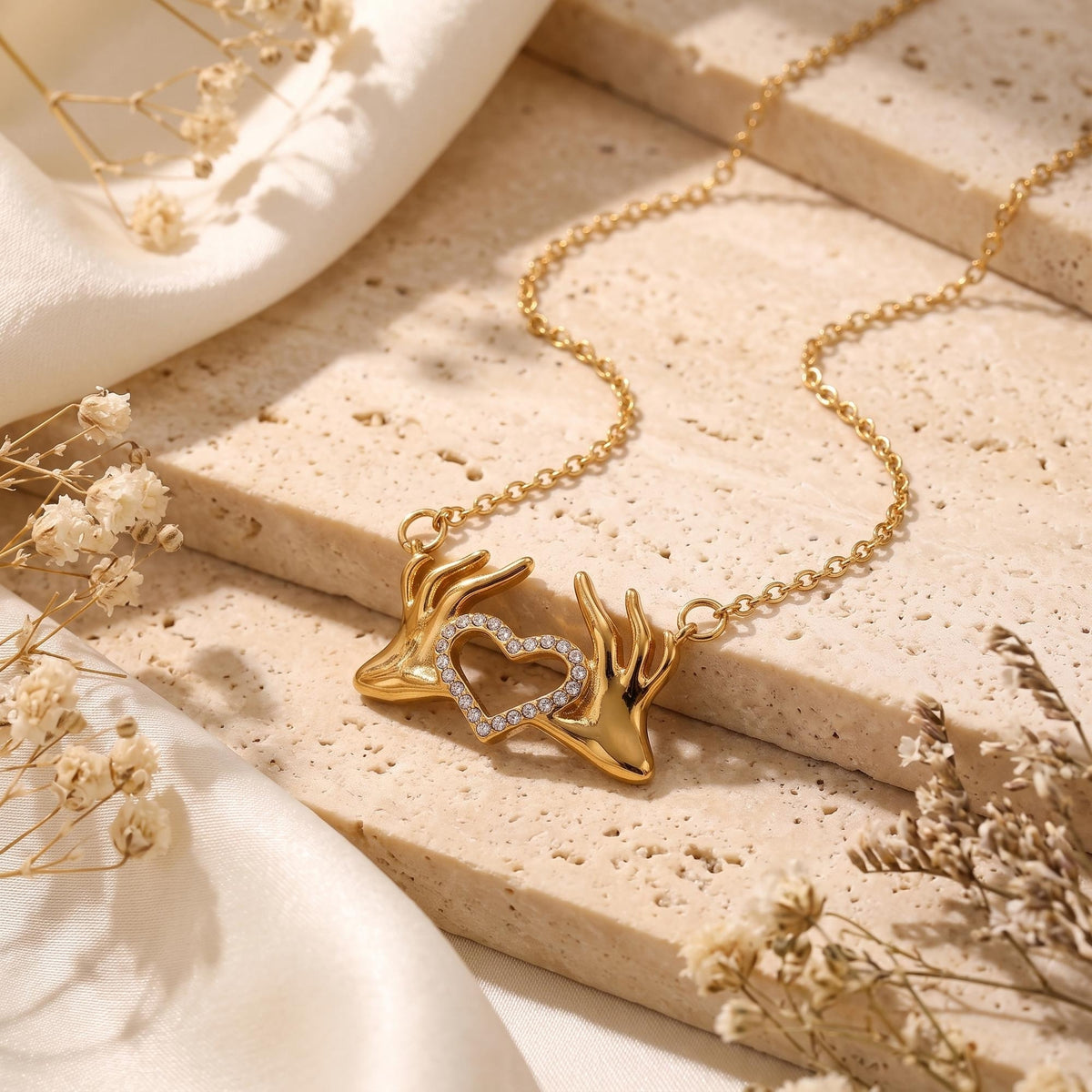Gold-tone heart hands pendant necklace with clear cubic zirconia, displayed on travertine with silk and dried flowers.
