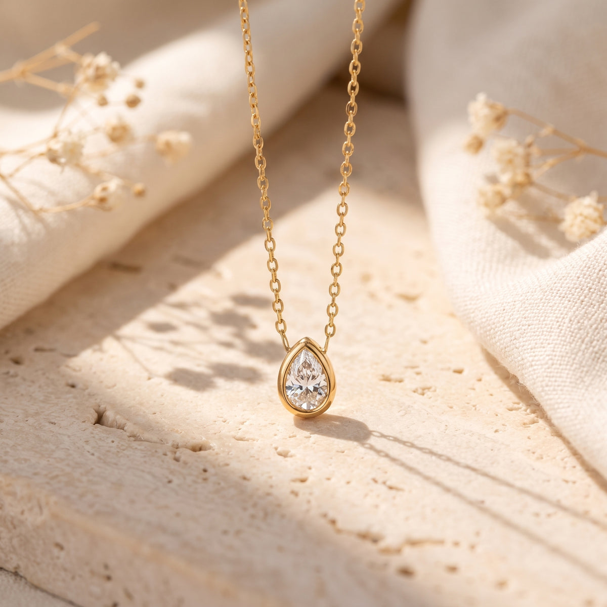 Gold-tone bezel necklace with a pear-cut clear stone on a stone surface with soft cream fabric.