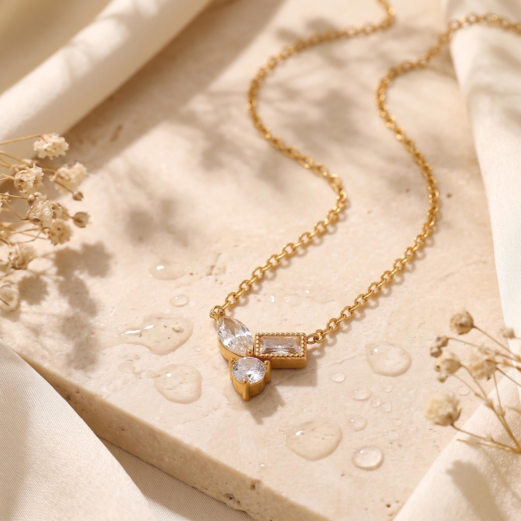 18K gold geometric necklace with marquise and baguette cubic zirconia on a stone slab with water droplets.