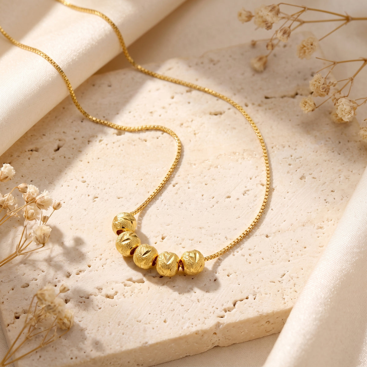 Gold-tone box chain necklace with five carved beads laid on a light beige stone slab among dried flowers.