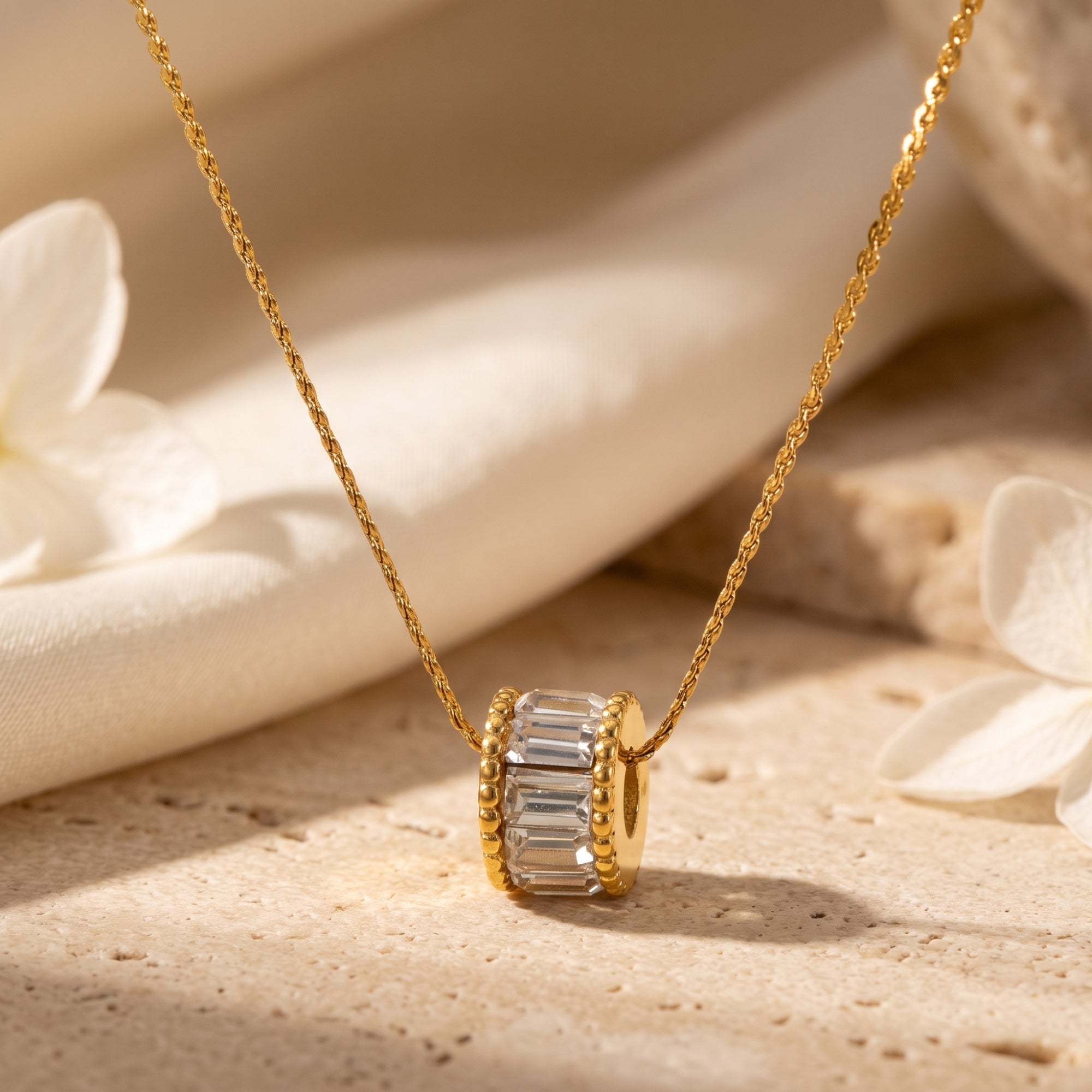 Gold pendant necklace with baguette-cut simulated diamonds on a stone surface with white flowers.