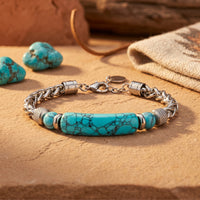 Silver stainless steel wheat chain bracelet with a turquoise-hued focal bar on a desert-themed stone surface.