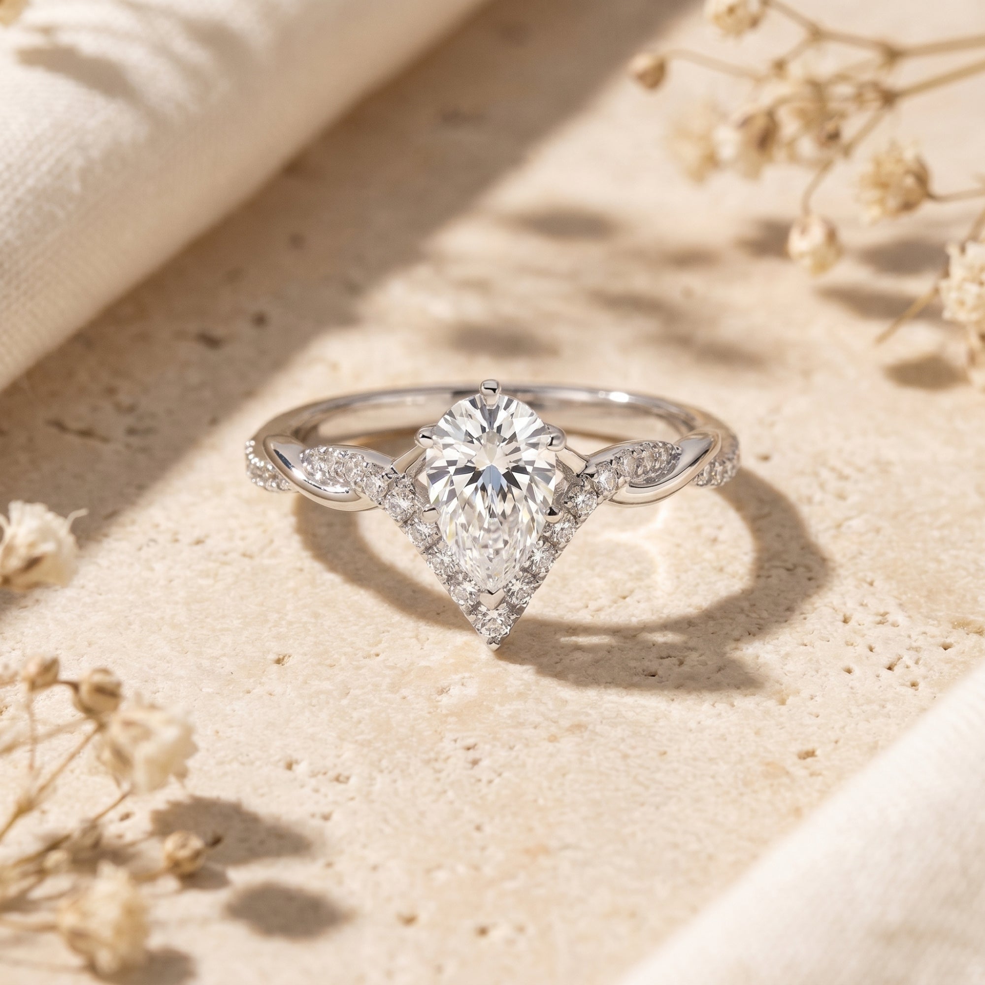 Silver pear-cut moissanite promise ring with a chevron halo and twisted band on a stone surface.