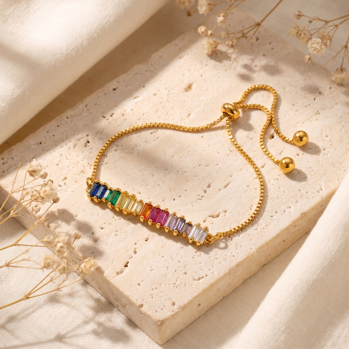 Gold-tone adjustable slider bracelet with a bar of colorful rainbow baguette stones on a cream stone slab.