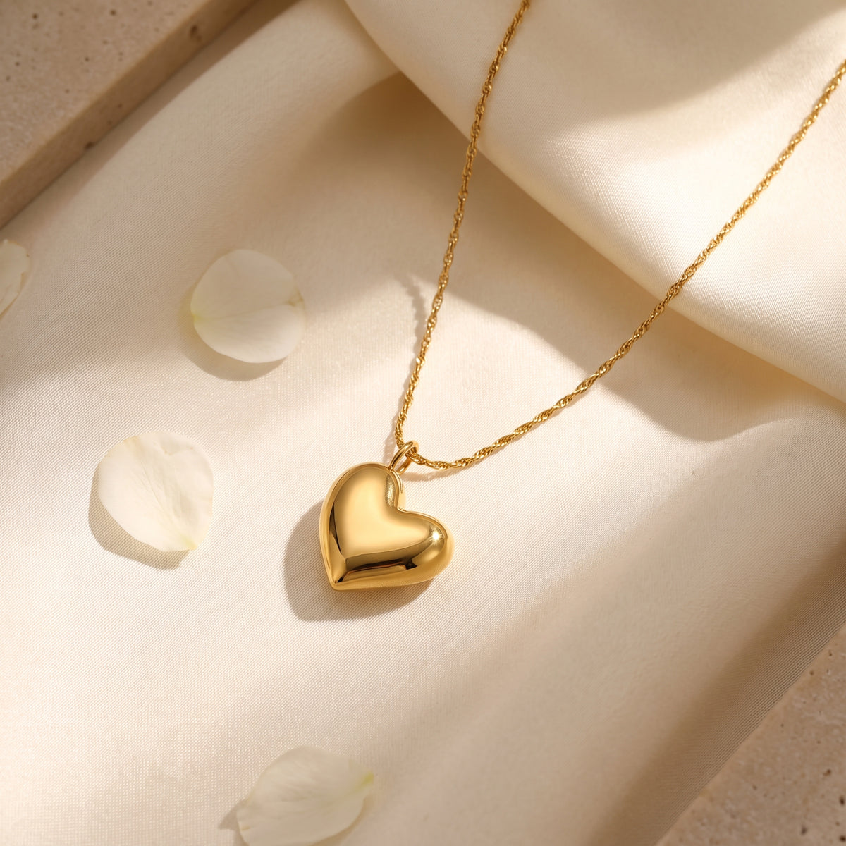 Close-up of a gold-tone puffed heart necklace on light fabric with white flower petals.