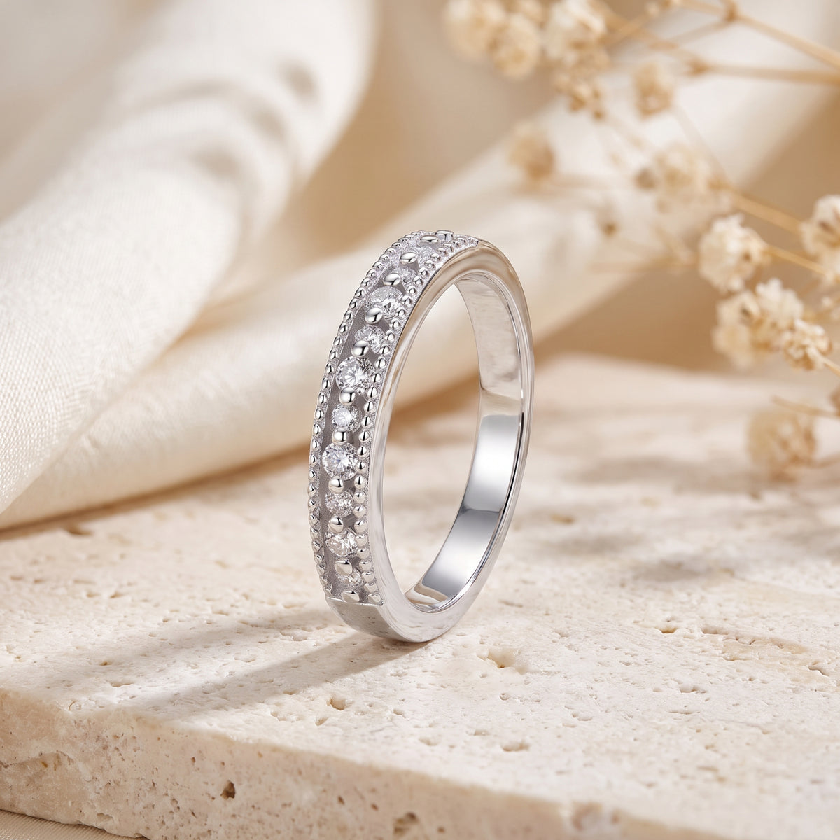 Silver band with bead-set clear stones and milgrain edges standing upright on a textured surface.